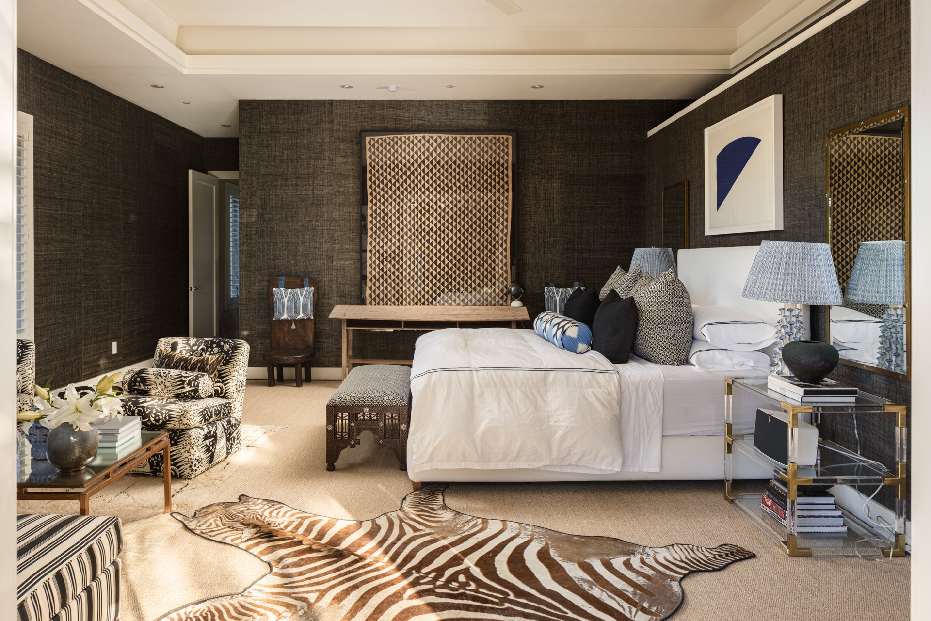 Luxurious bedroom with a white bed, black and white patterned armchair, and zebra rug, dark textured walls, modern artwork, nightstands with lamps, and decorative pillows and furniture.