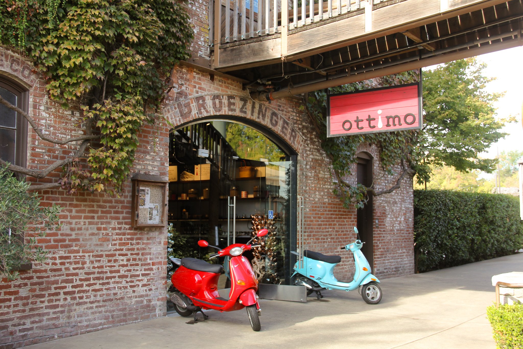 Exterior view of a brick building with a sign that reads 'Ottimo'. There are two scooters, one red and one blue, parked outside near the entrance, and some greenery and trees in the background.