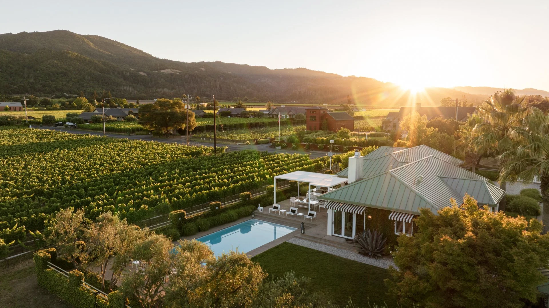 Sunset over a suburban backyard with a pool, patio, and lush greenery, overlooking fields and mountains in the distance.