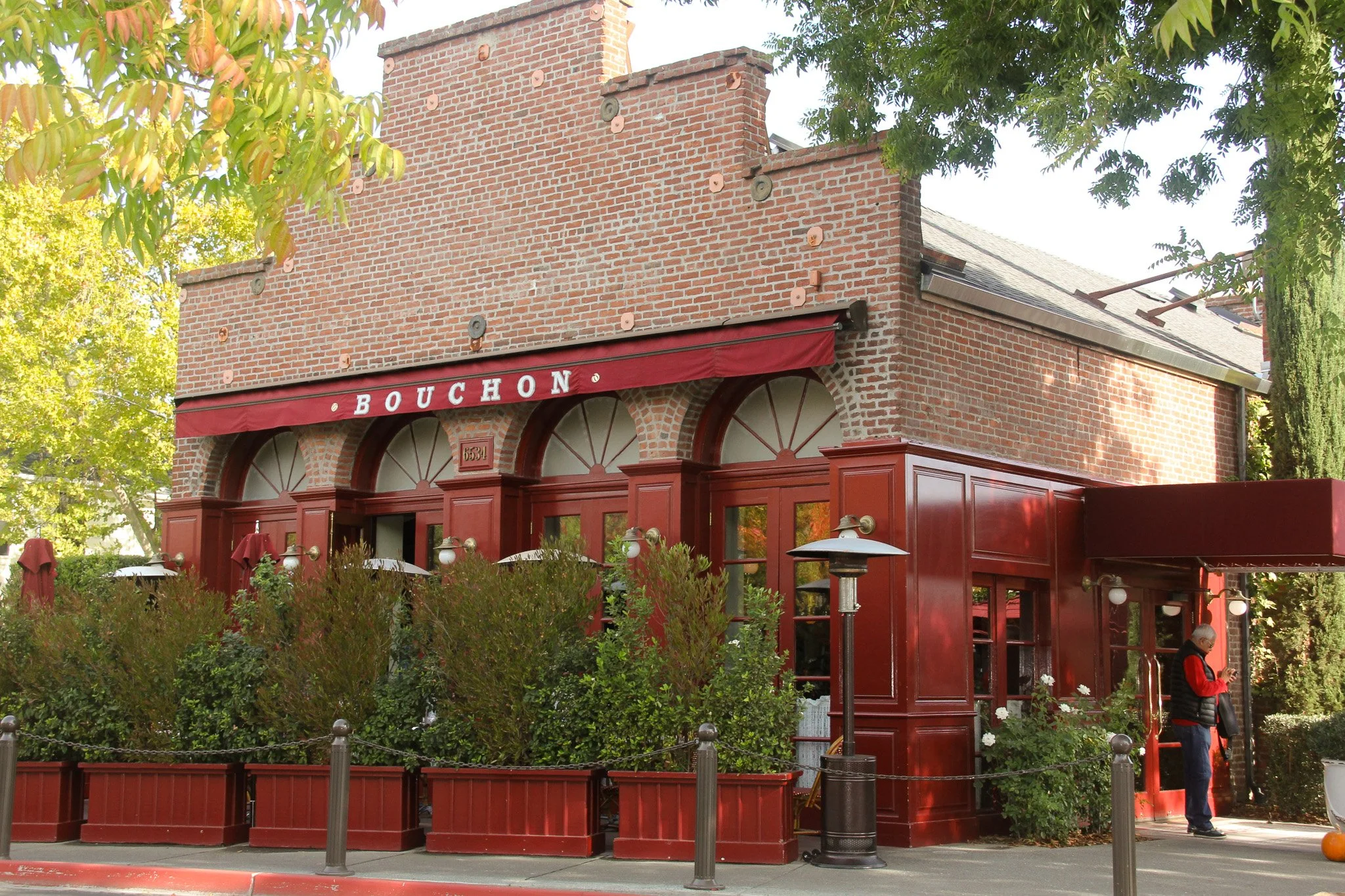 A vintage red brick building with arched windows and a red awning with the sign 'BOUCHON'. There are green trees surrounding the building, and a person is standing on the sidewalk near the building.