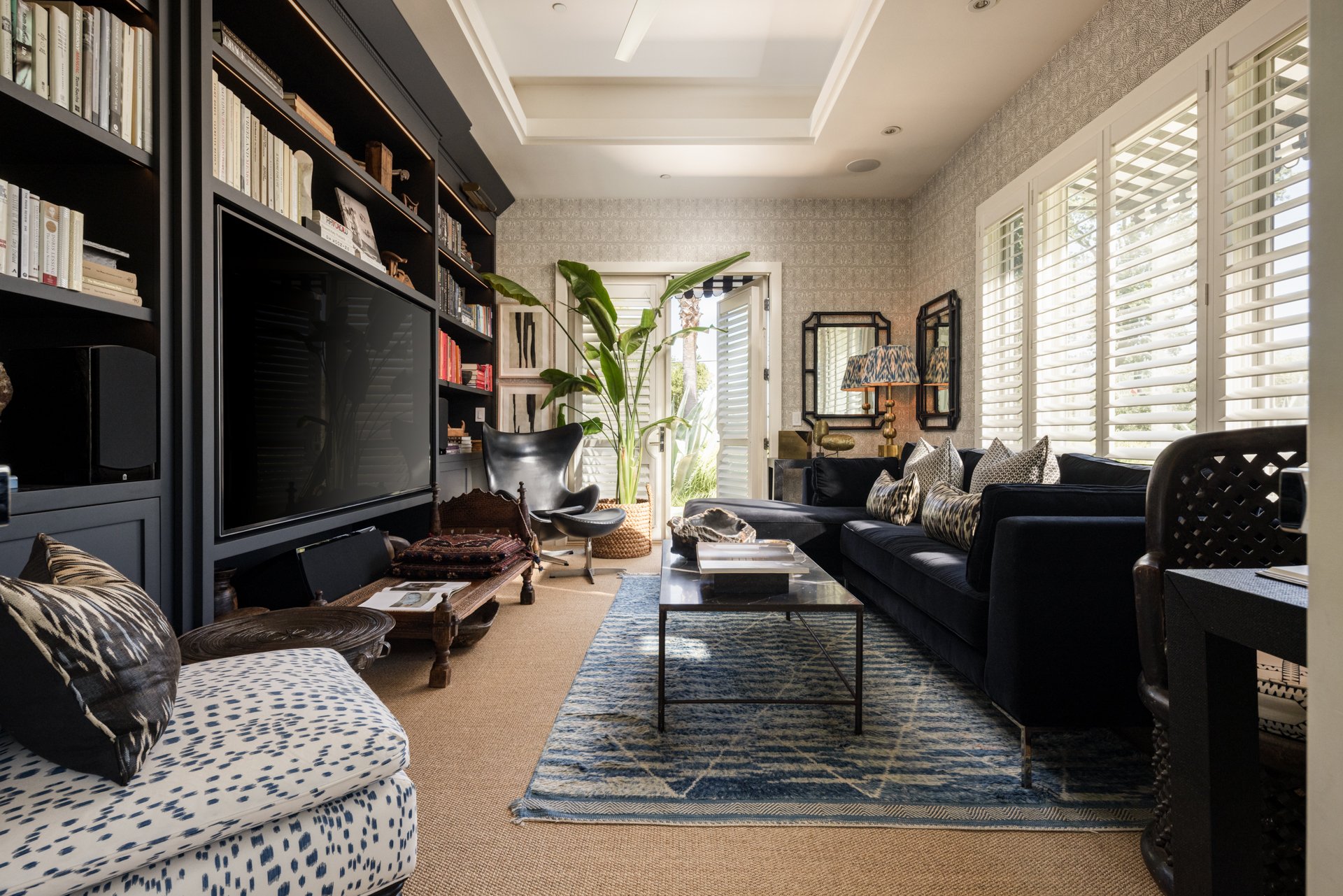 A living room with a black sofa, patterned pillows, a glass coffee table, large windows with white shutters, a tall plant, framed art, a marble lamp, and a black bookshelf filled with books and decorations.