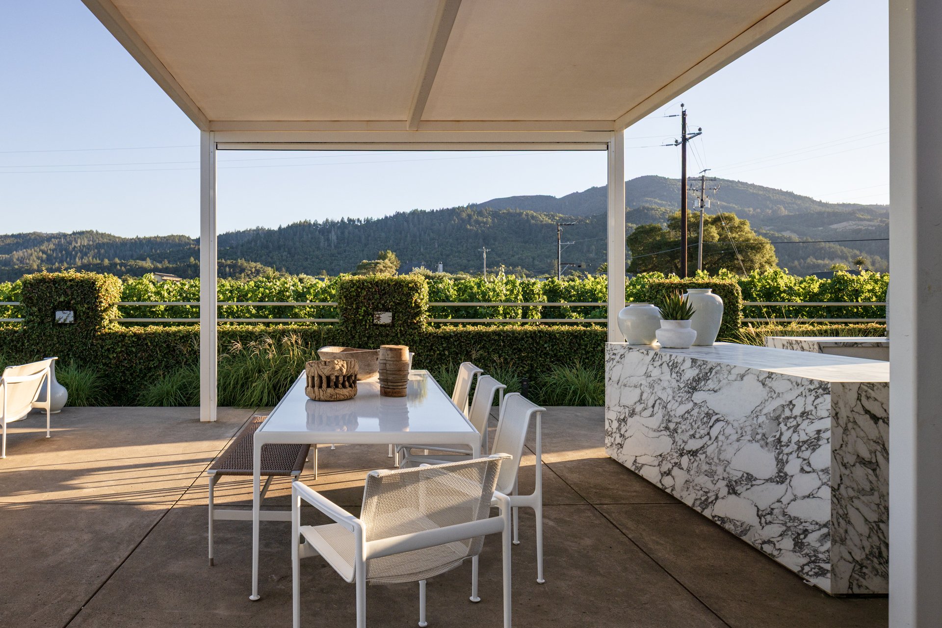 Outdoor patio area with white dining table, white and brown chairs, decorative vases on a marble counter, and lush green hills and mountains in the background under a beige roof.