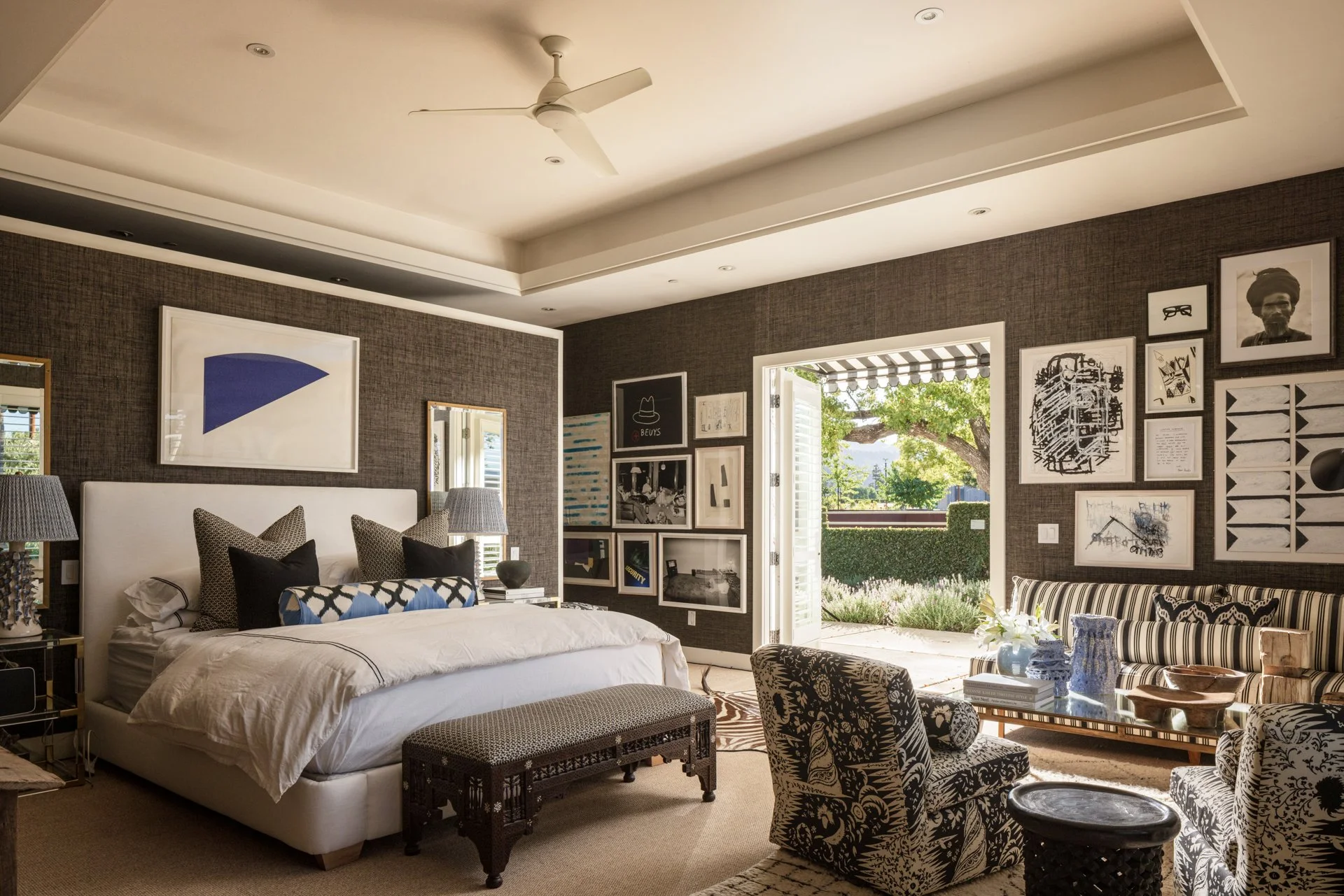 A bedroom with a bed, artwork on the walls, and an open door leading outside to a garden with trees and shrubs.