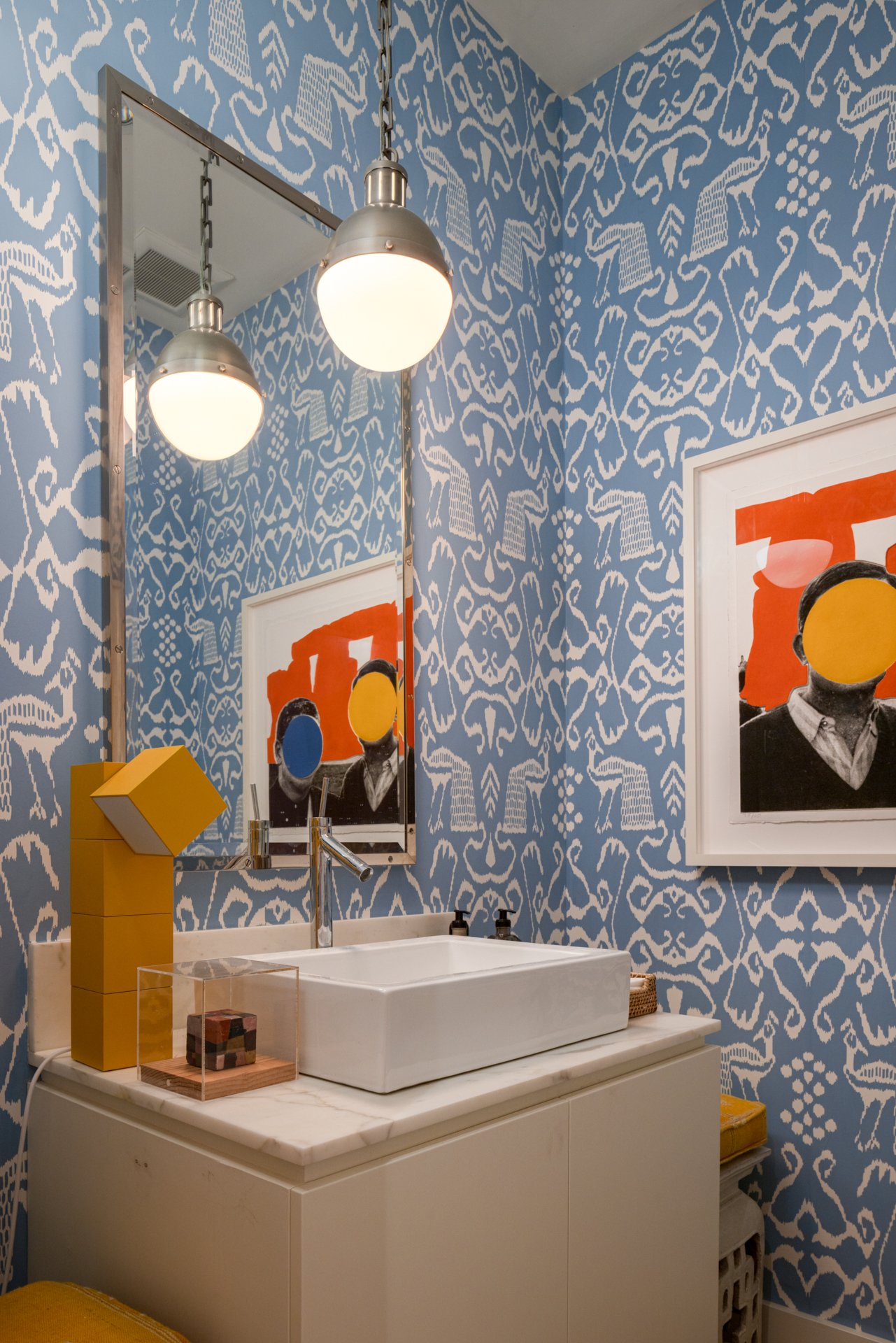 A bathroom with blue and white patterned wallpaper, a rectangular white sink, a yellow abstract sculpture, and framed artwork with a person holding two colored circles over their face.