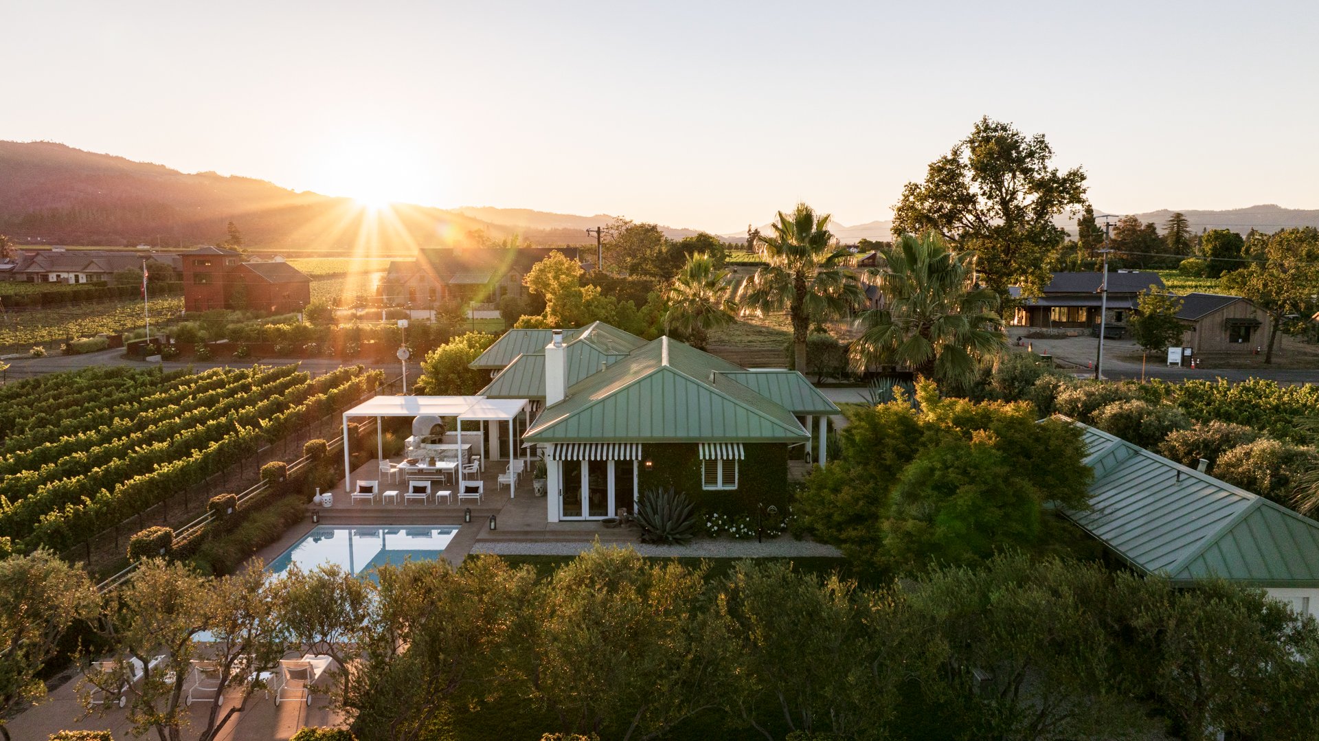 Sunset over a scenic landscape with a house, swimming pool, surrounding trees, mountains in the background, and a vineyard.