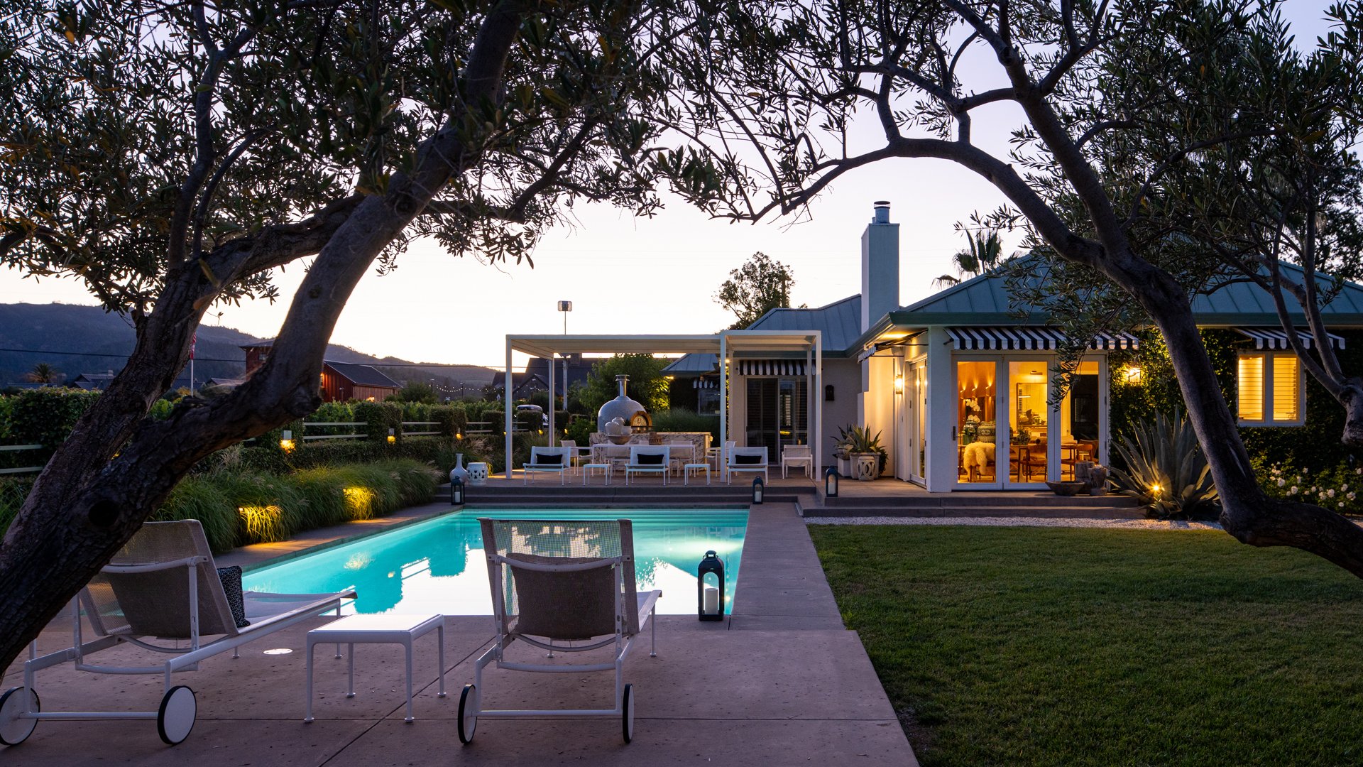 A backyard with a swimming pool, outdoor seating area, and a house illuminated at dusk. Trees frame the scene.