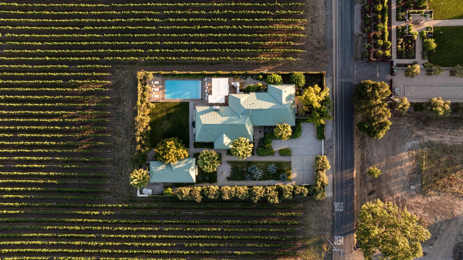 An aerial view of a house with a landscaped yard, swimming pool, and neighboring rows of vineyards, with a road on the right side.