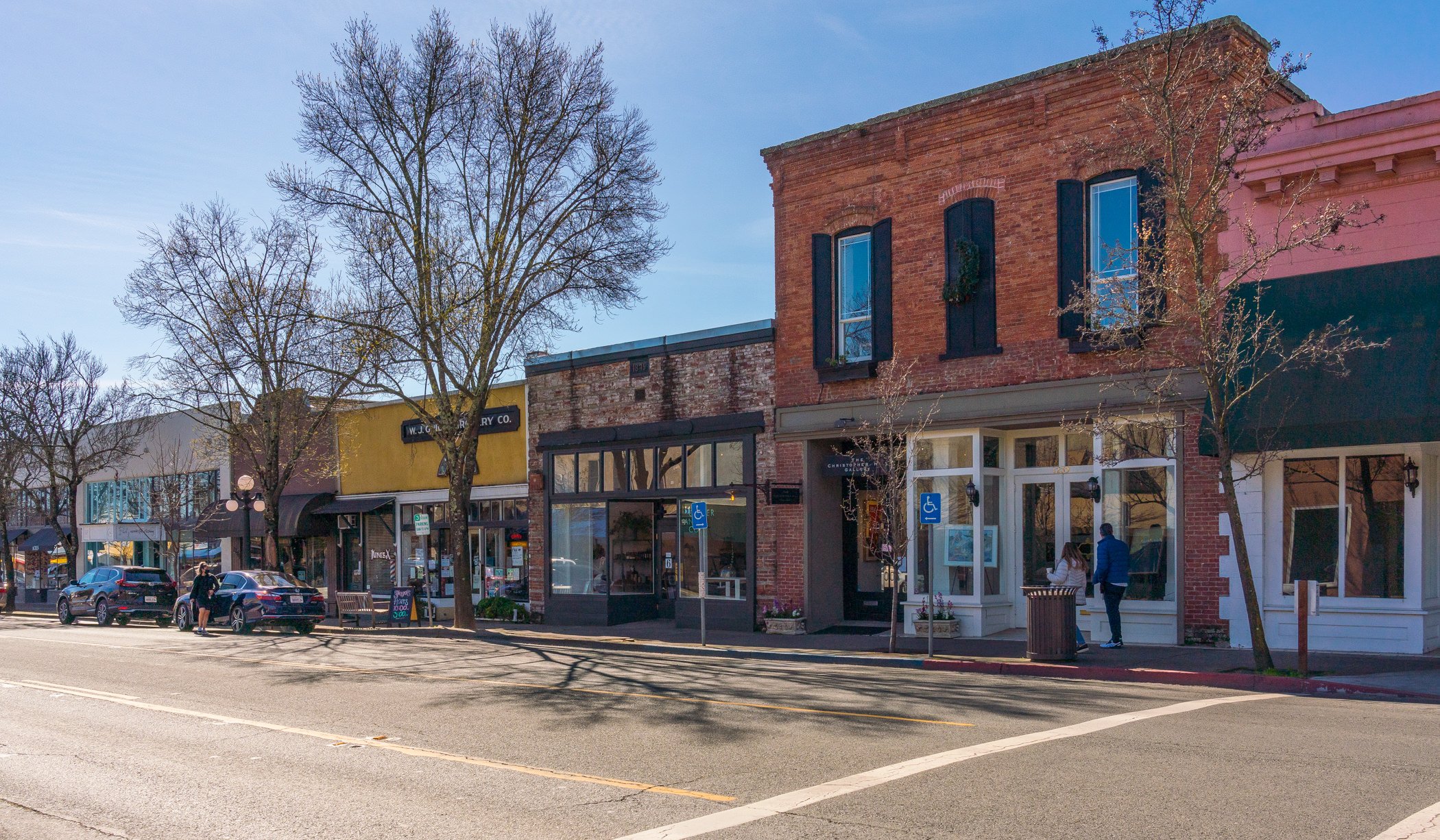 A city street with storefronts, trees, and parked cars. Two pedestrians are walking on the sidewalk, and the buildings are a mix of brick and painted facades.