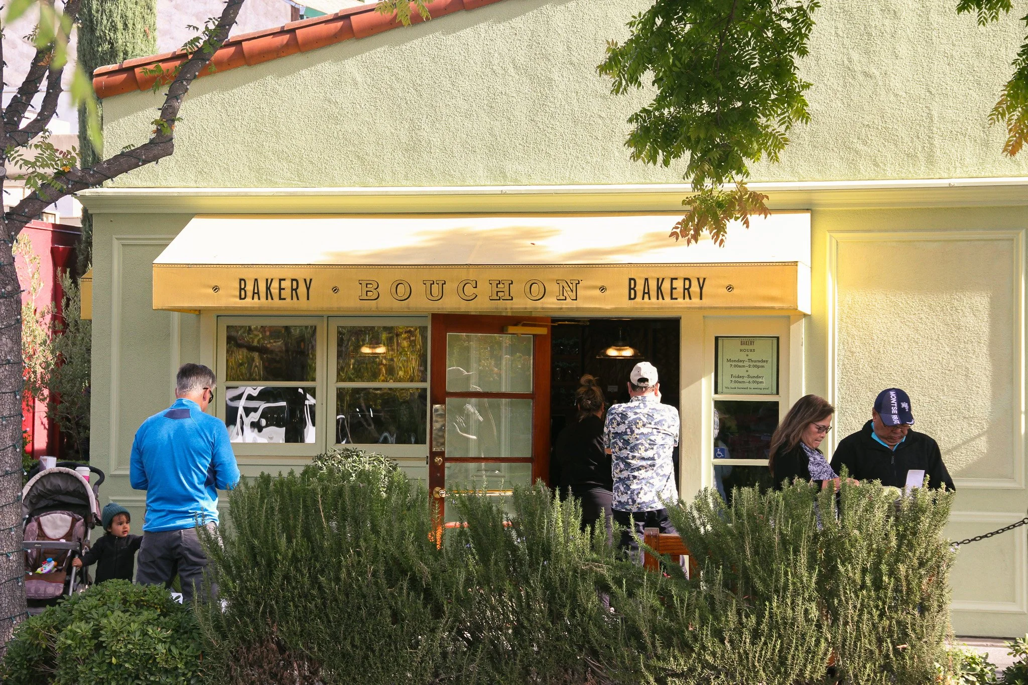 Front view of a bakery named 'Bouchon Bakery' with several people standing outside, some looking at the bakery and others engaging in conversations. There are trees and greenery in front of the bakery and a sign with bakery hours on the right side of