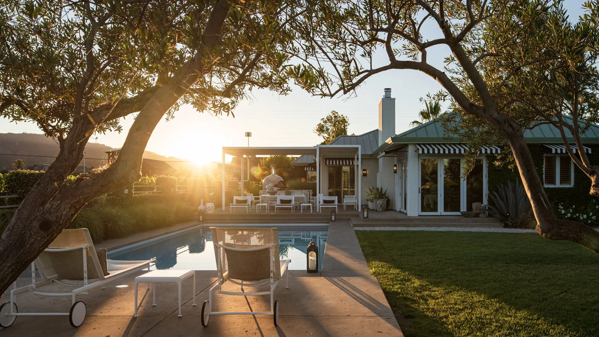 Sunset view of a backyard with a swimming pool, white outdoor furniture, and a patio area with a house featuring a chimney. Large trees frame the scene.