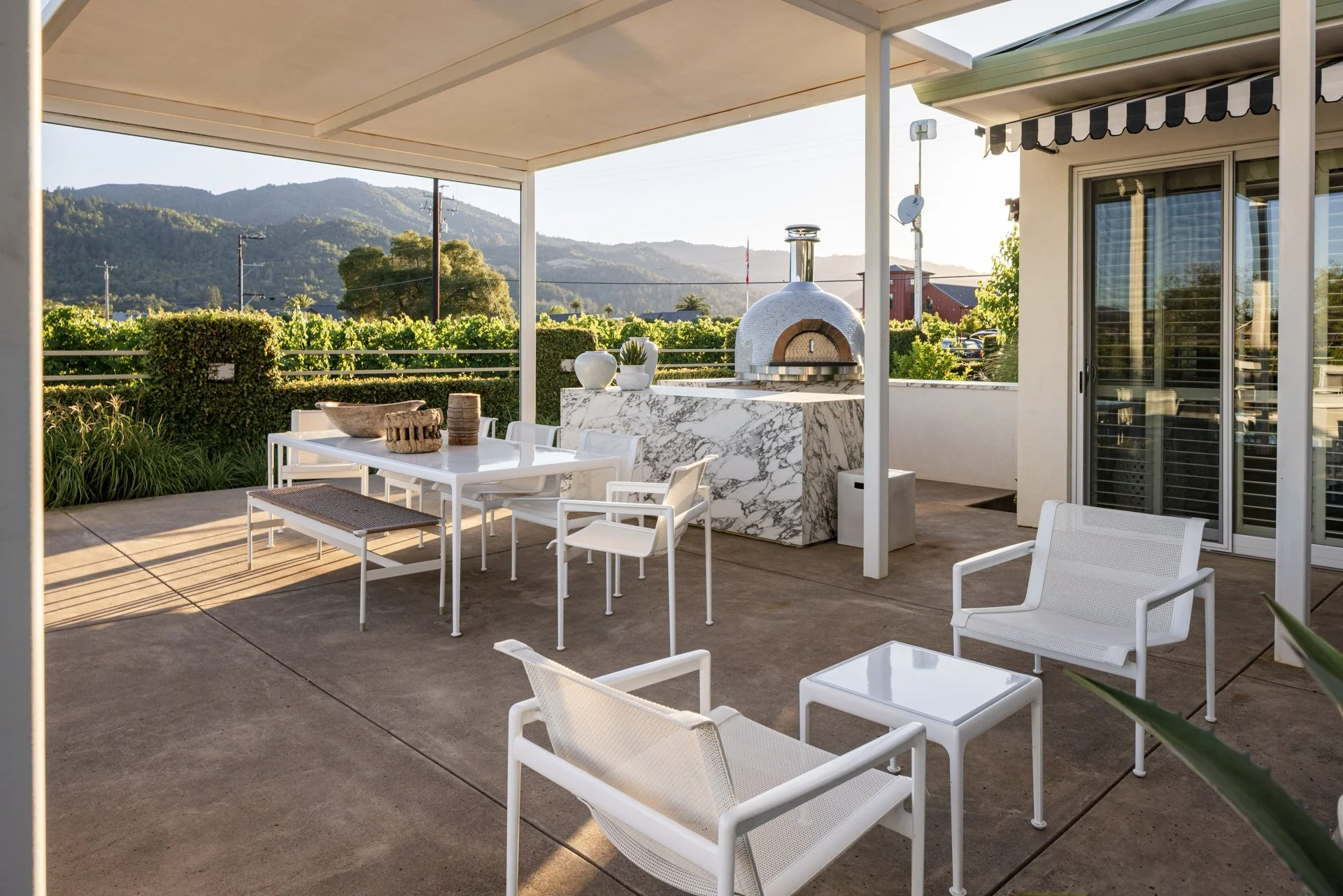 Outdoor patio area with white furniture, a marble pizza oven, and mountain view in the background.