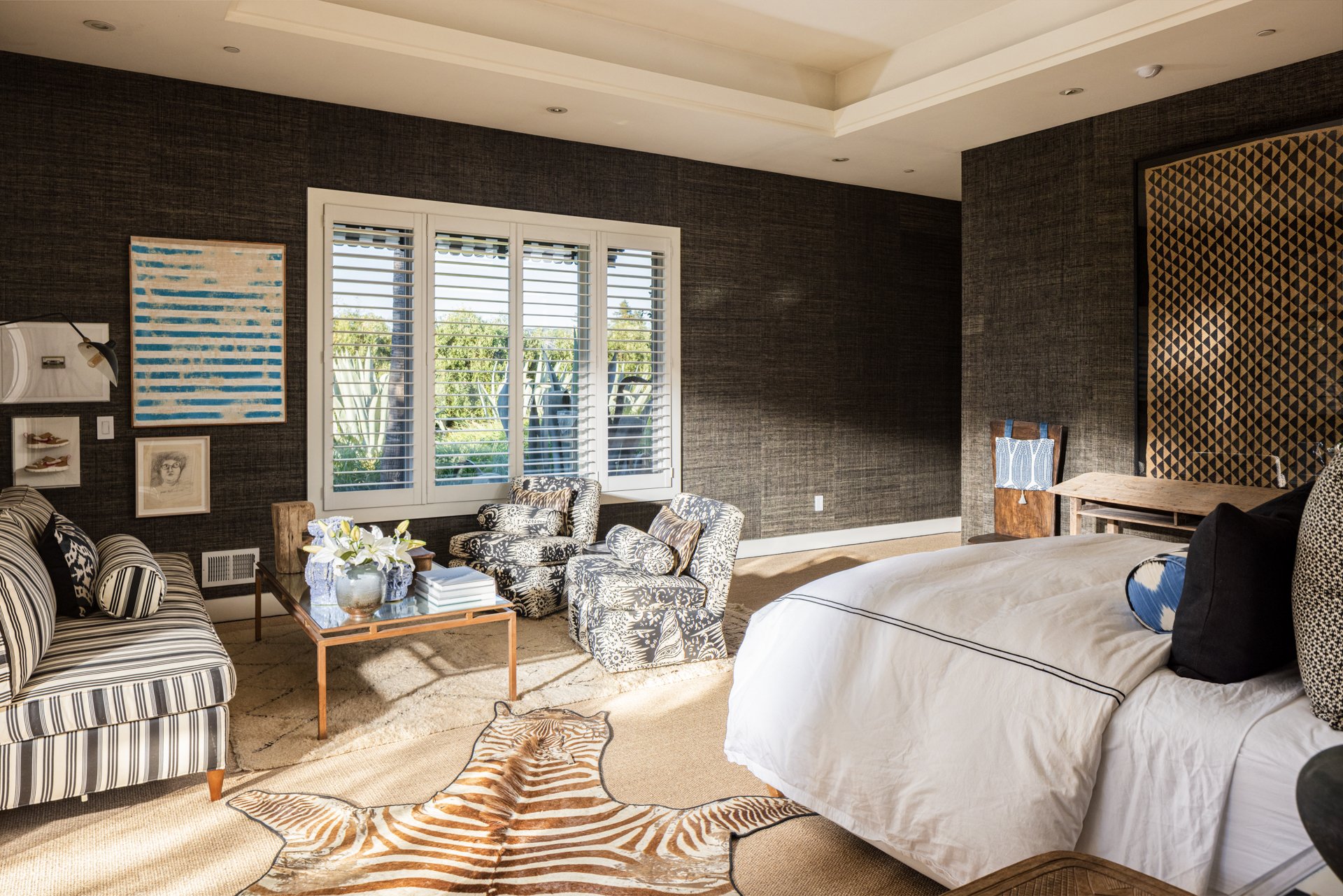 A spacious hotel room with a bed, a sitting area with chairs and a coffee table, a large window with blinds, and artwork on the walls. The room has dark textured walls and a patterned rug.