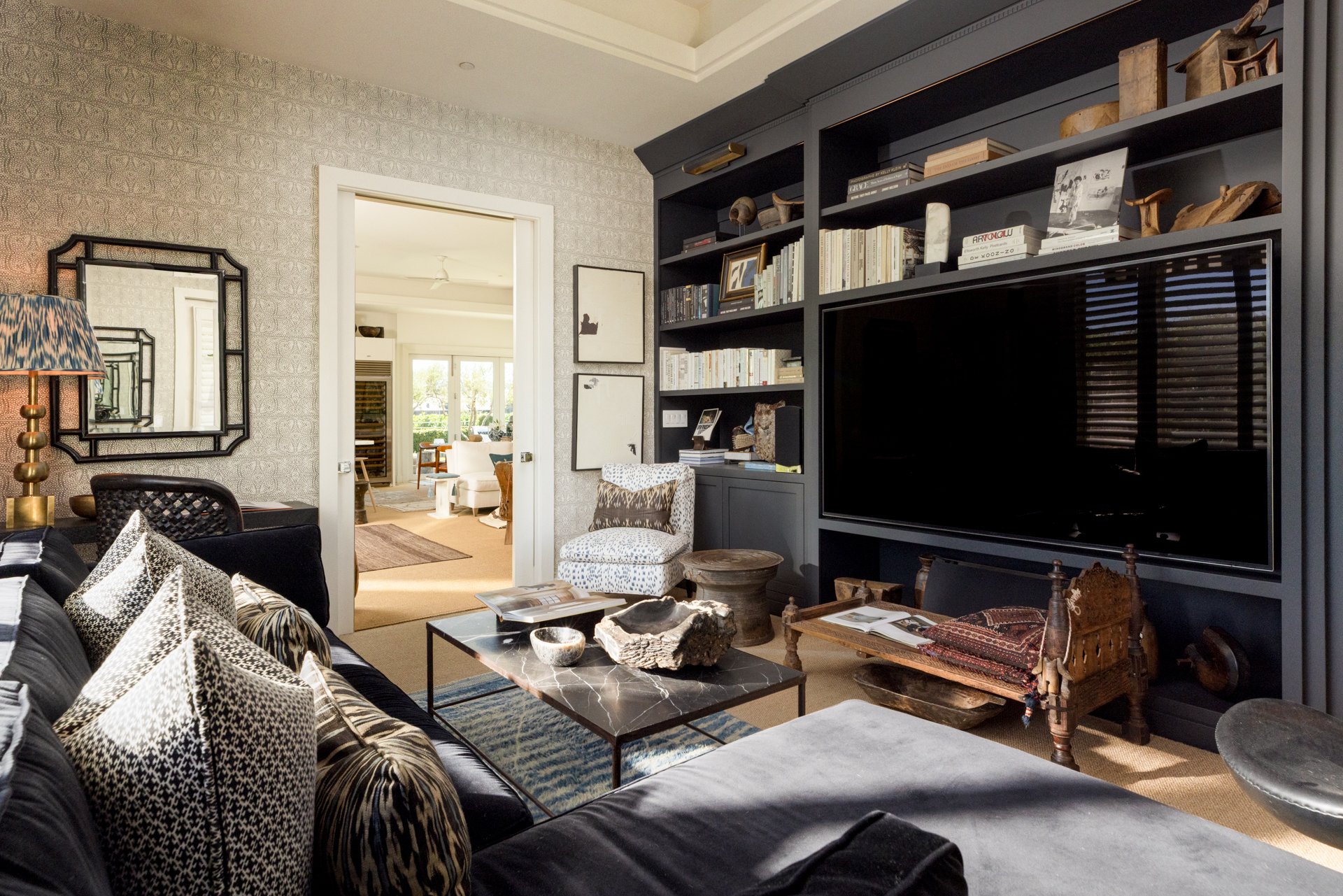 Living room with black sofa, patterned pillows, and coffee table with decorative items. Dark bookshelf with books and decorations, leading to a bright adjoining room with armchair and large windows.
