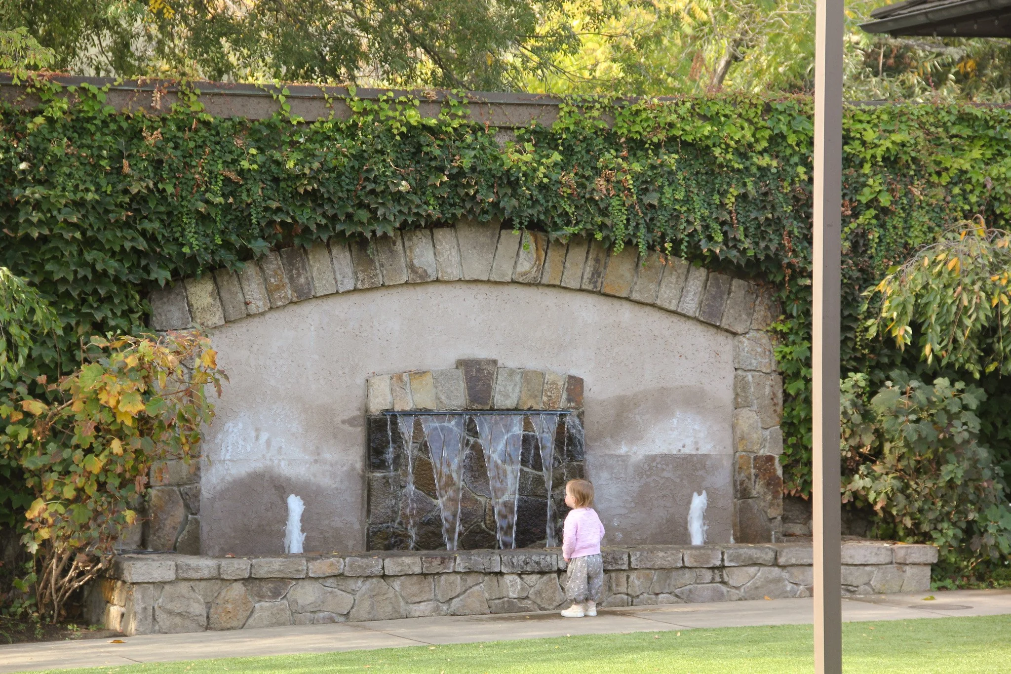 A stone wall with an arched water fountain, surrounded by green ivy and trees, with a little girl in a pink shirt and white pants standing in front of it.
