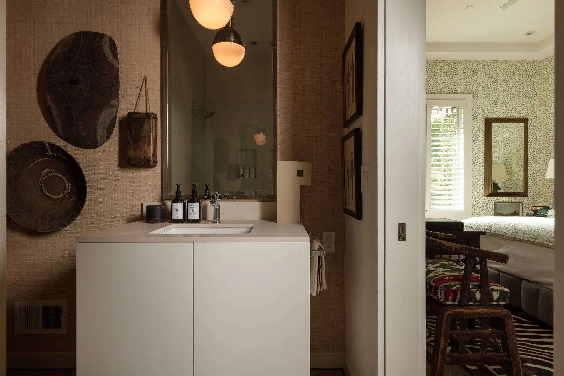 A small bathroom with a white vanity, a mirror, and wall-mounted art, adjacent to a bedroom with a patterned wall, a window with blinds, and a bed with a lamp and artwork.