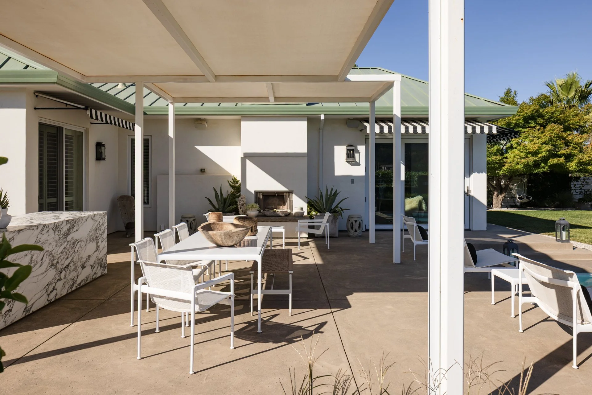 Patio area with white outdoor furniture, a marble-topped bar, and a fireplace, enclosed by a pergola with a green roof on a sunny day, with trees and a swing in the background.