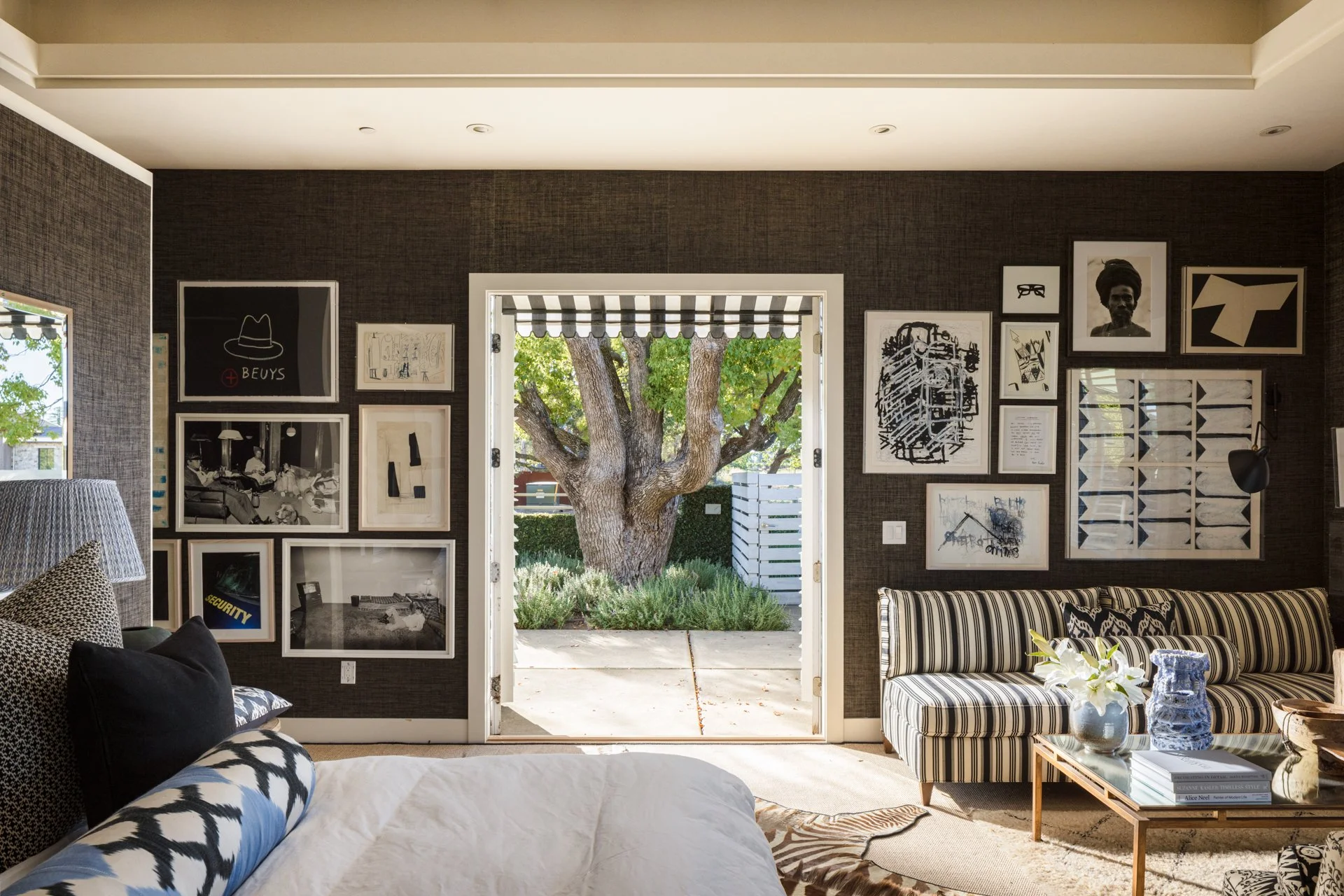 Living room with dark textured walls and a gallery wall featuring various framed artwork and photographs, including a black-and-white portrait of a woman. An open doorway shows a view of a garden with a large tree and greenery outside. Interior decor