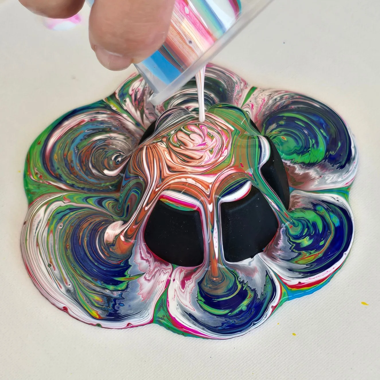 Colorful swirling paint in vibrant colors like pink, green, blue, and orange on a white surface, with a hand pouring more paint into the center.