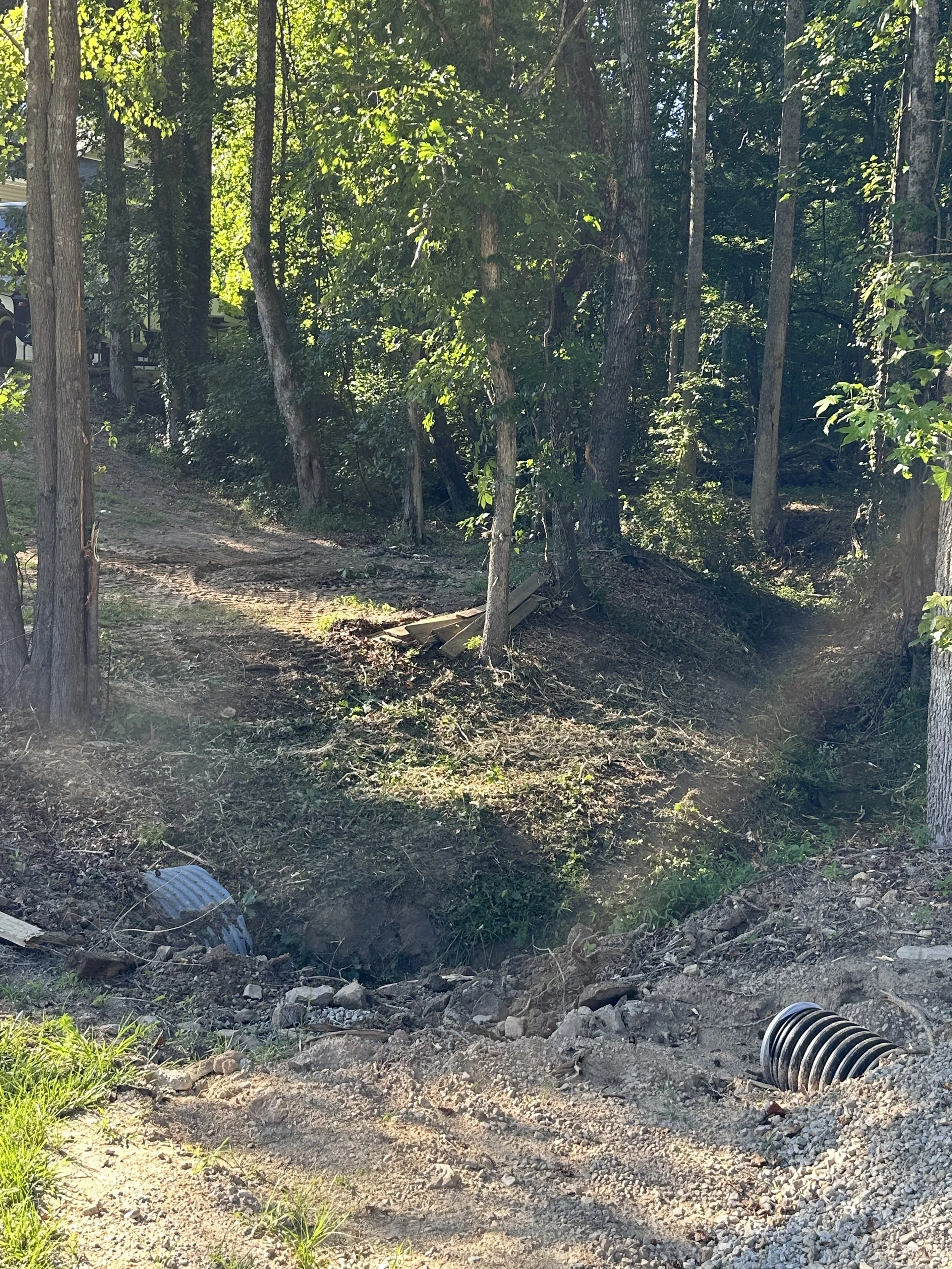 A wooded area on a creek bed with trees and a dirt path. There are construction materials, including a large black corrugated pipe.