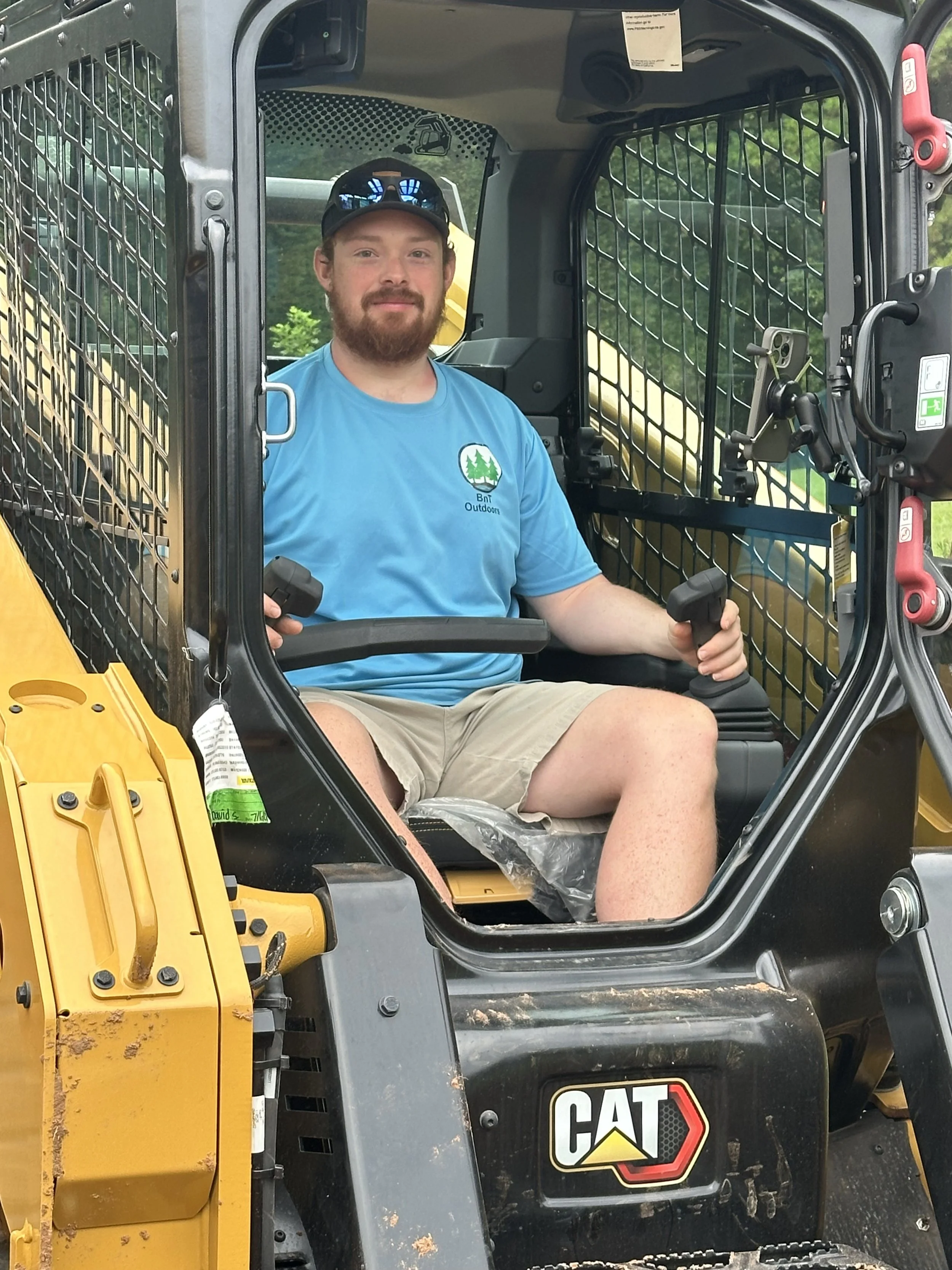 A man with a beard and sunglasses on his head sitting in the driver seat of a yellow and black CAT construction vehicle, holding the controls with both hands. He is wearing a blue T-shirt with a logo that says 'Bnt Outdoor' and shorts. The vehicle has protective metal bars around the cabin and a partially visible yellow attachment in the background.