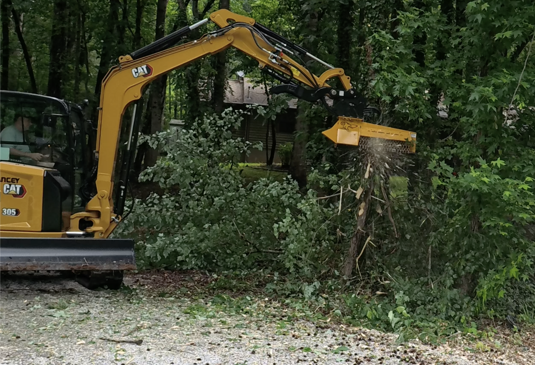 A yellow Caterpillar backhoe with a front loader is clearing trees and bushes in a wooded area. Debris and cut branches are scattered on the ground.