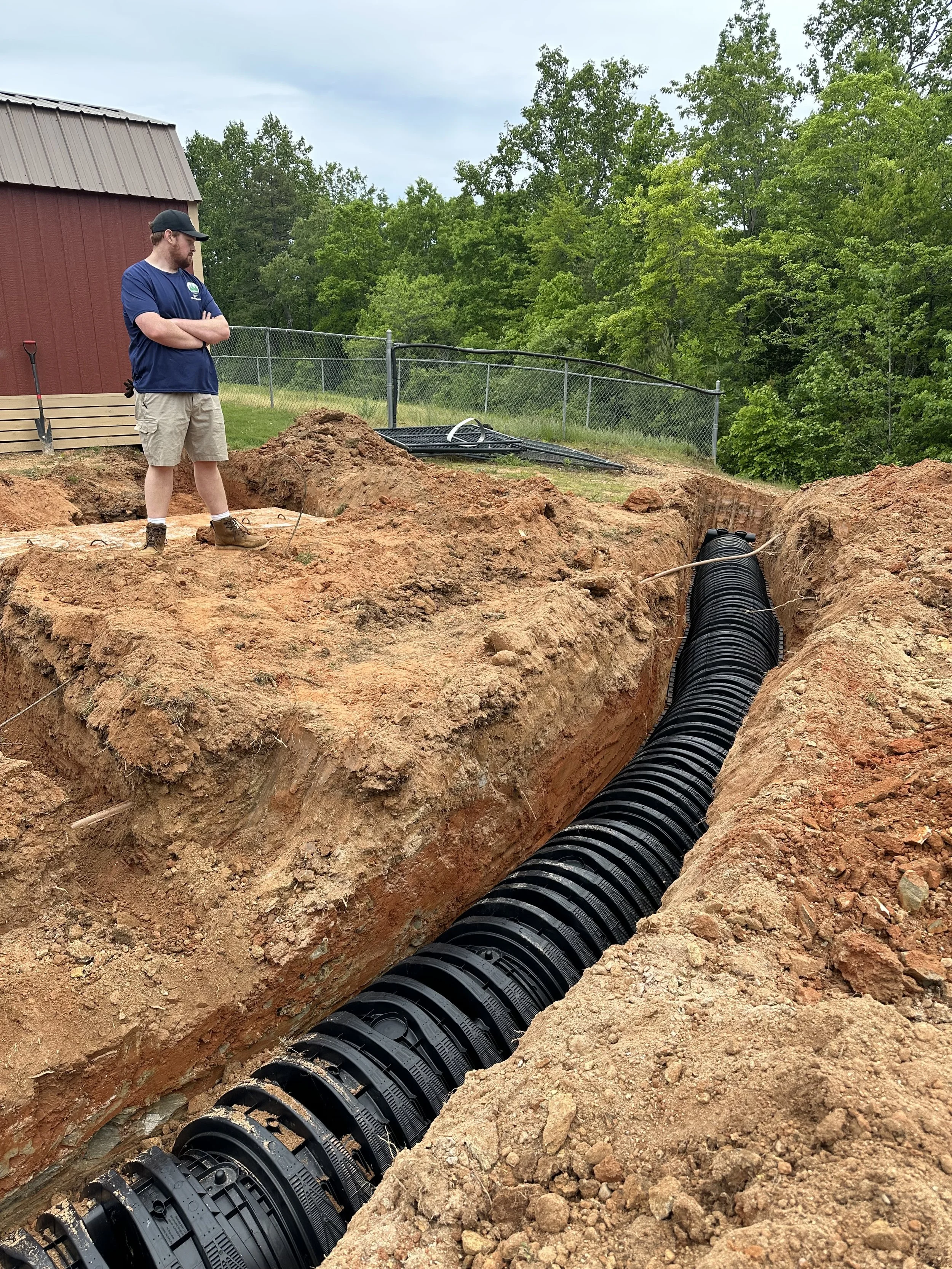 A person standing near a large trench with a black corrugated pipe underground, clean dirt around it, and trees and a red barn in the background.