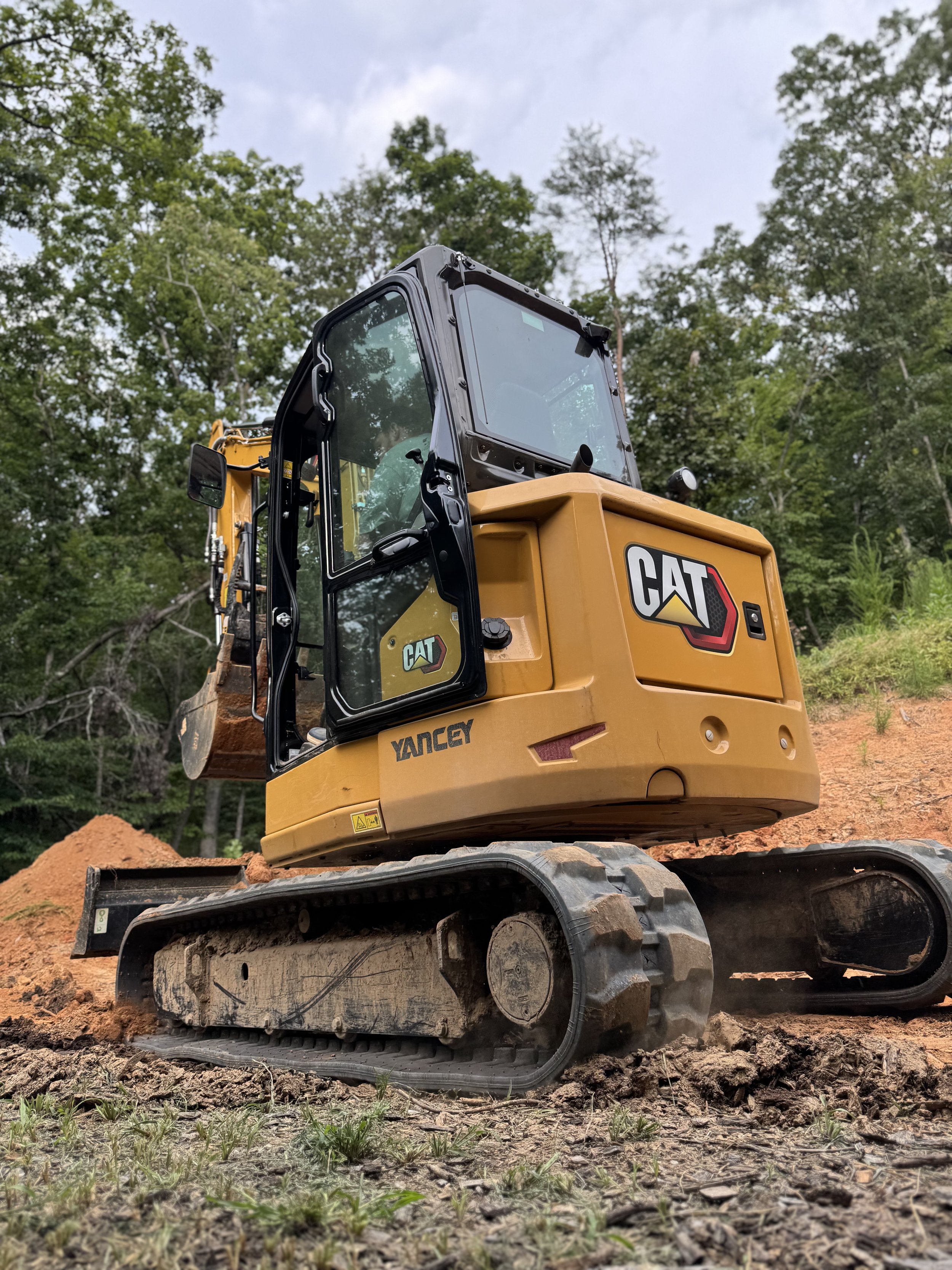 A yellow Caterpillar excavator with a black cabin and tracks, working outdoors on dirt and surrounded by green trees, under a cloudy sky.