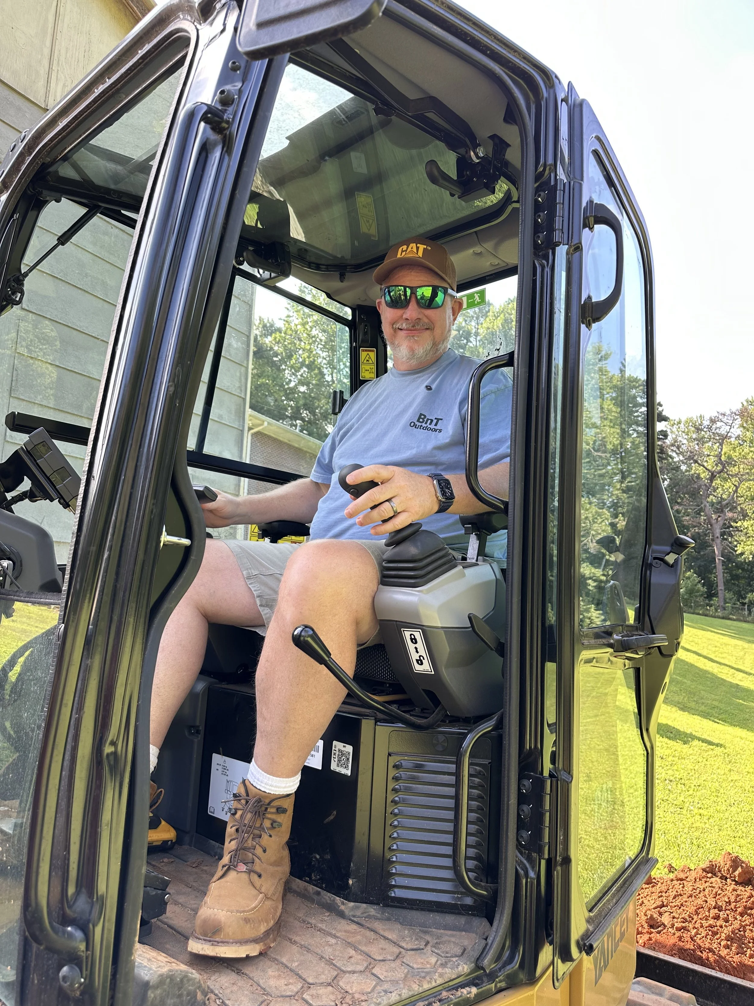 A man sitting in the operator's seat of a compact excavator, smiling, wearing sunglasses, a baseball cap, a T-shirt, shorts, and work boots. The excavator is outdoors near a house with trees in the background.