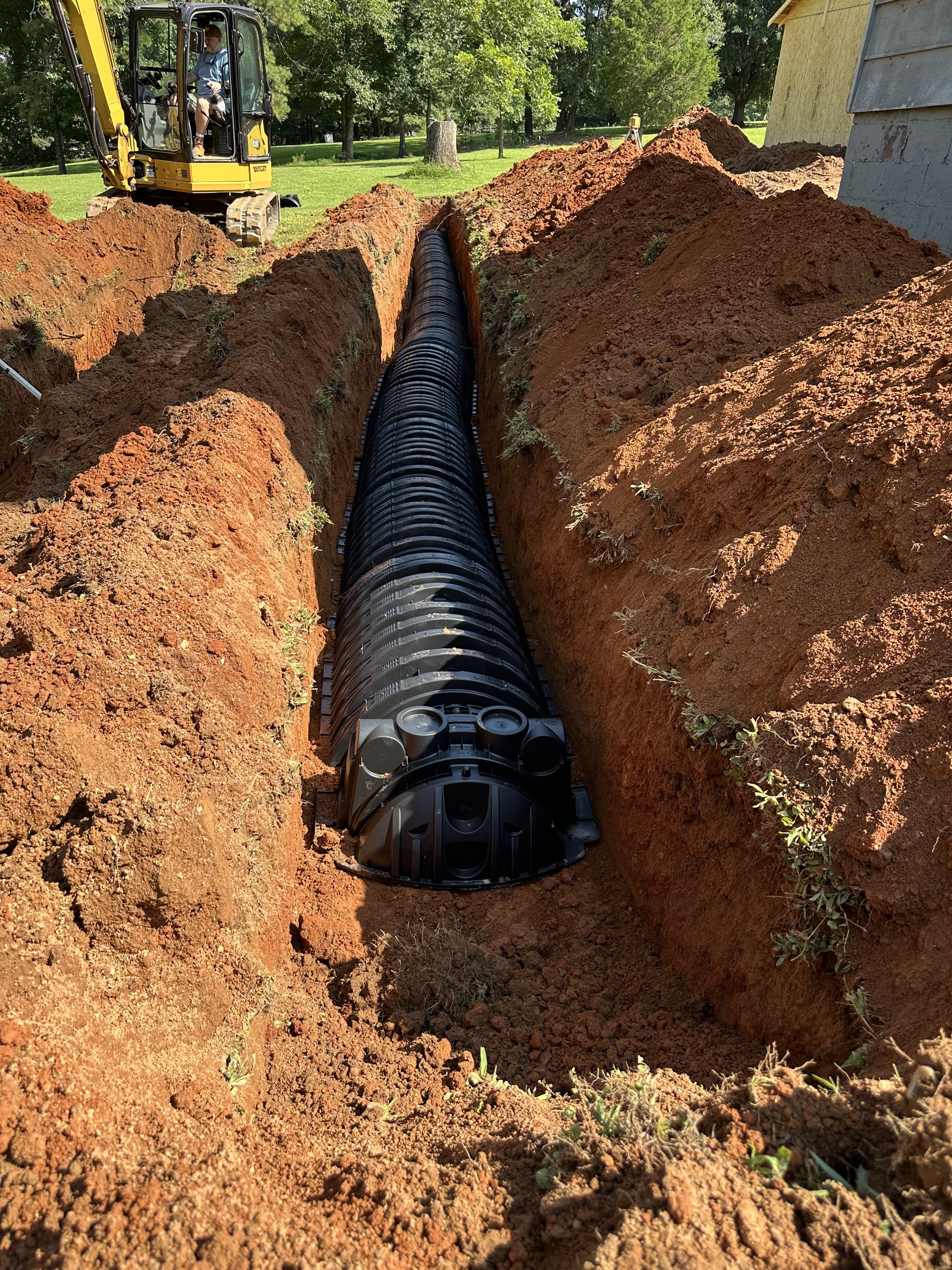 A construction scene with a yellow excavator operating next to a house, digging a trench for underground piping. The trench contains a large black plastic pipe and is surrounded by piles of reddish-brown dirt and some green grass and small plants.