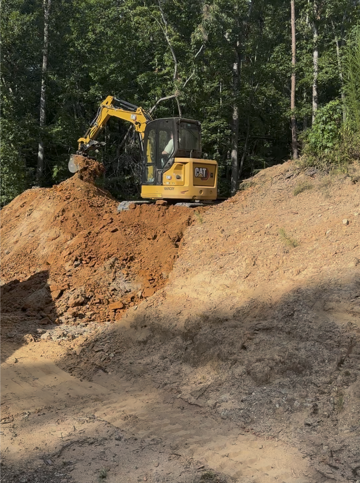A yellow and black CAT mini excavator working on a dirt hill with a dense forest of green trees in the background.