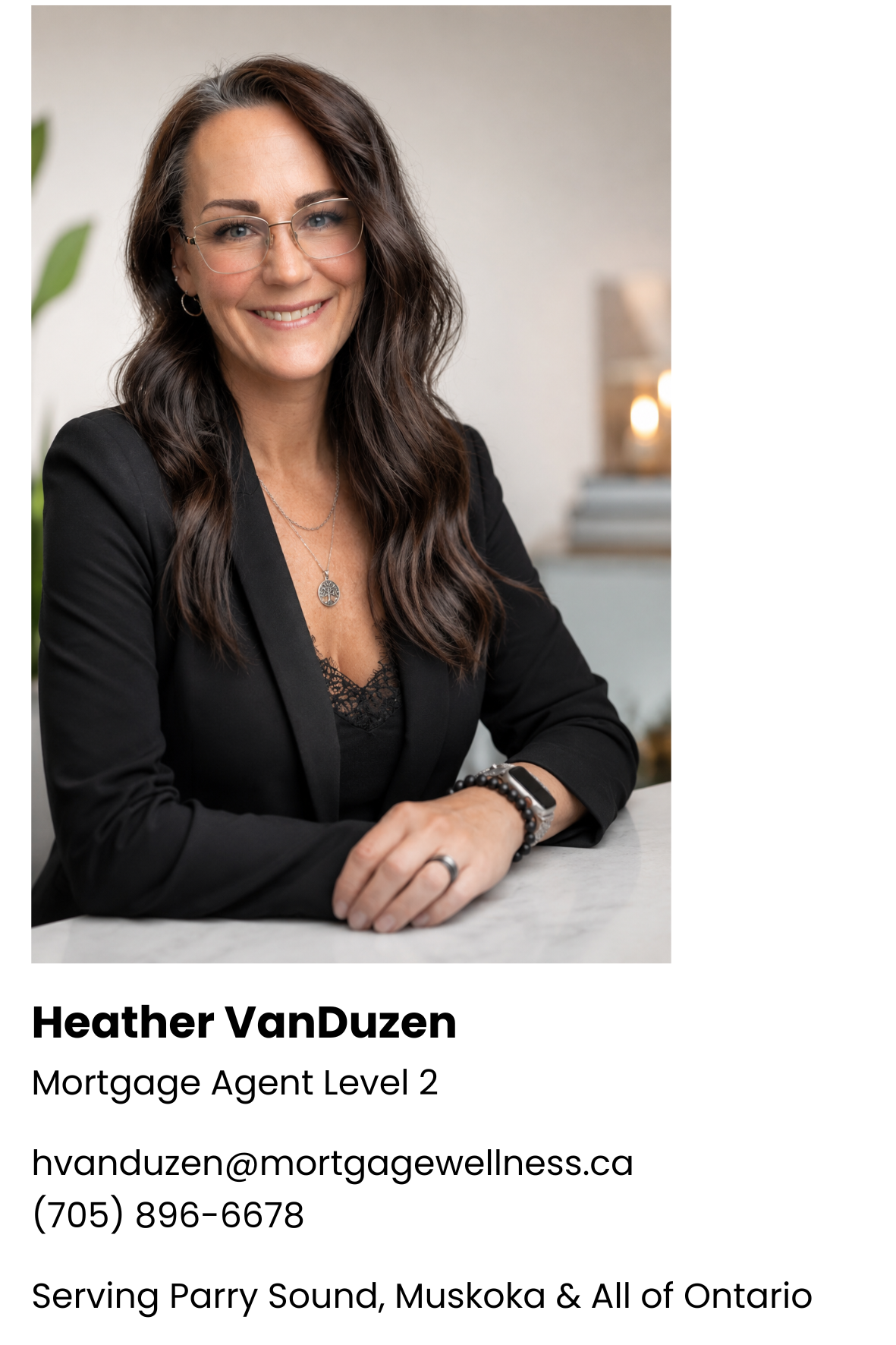 Headshot of Heather VanDuzen smiling, wearing glasses, a black blazer, with her hands folded on a white surface against a blurred background.