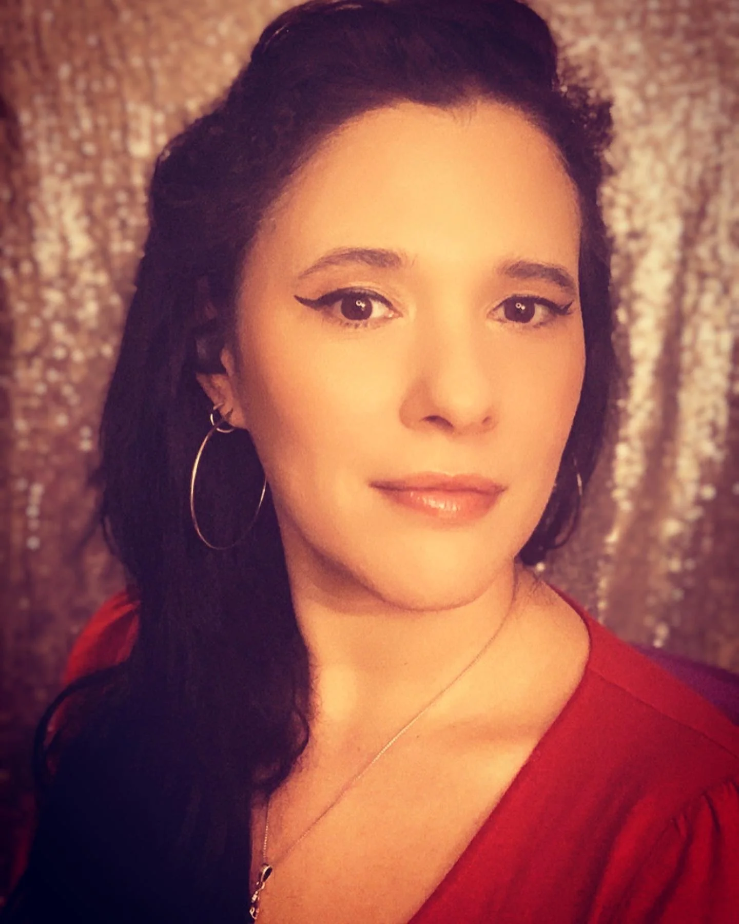 A woman with dark hair, hoop earrings, wearing a red top, smiling gently, in front of a textured, sparkly background.