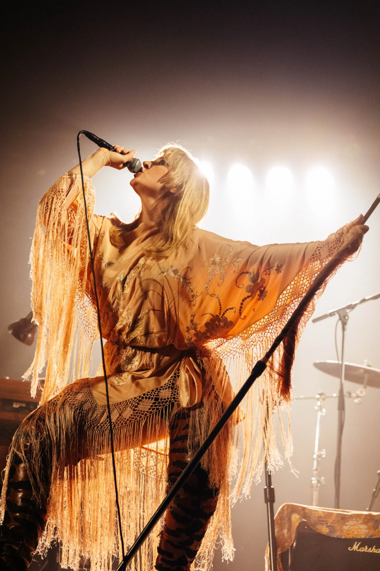 A female singer with blonde hair performs on stage, holding a microphone and wearing an orange embroidered shawl with fringes. She is backlit with stage lights, and musical equipment is visible in the background.