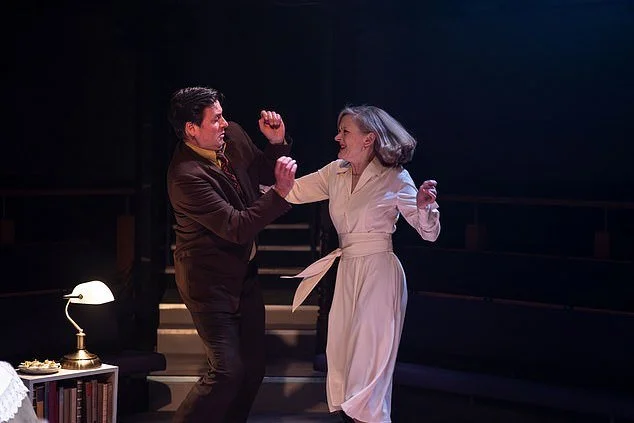 Two actors performing a dance or theatrical scene on stage, with a black background, a table with a lamp and books to the side, and stairs in the background.