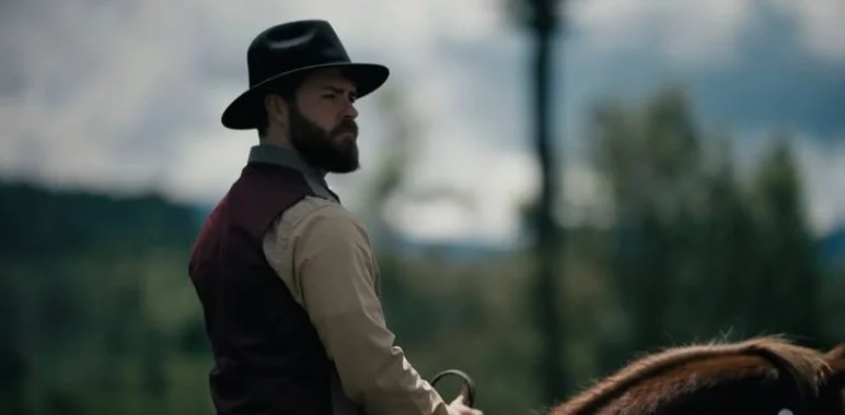 A man with a beard wearing a black hat, beige shirt, and dark vest riding a horse outdoors with a blurred background of trees and sky.