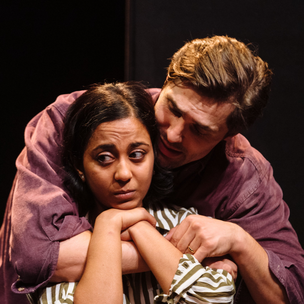Two actors performing on stage, one embracing the other, with a serious expression.