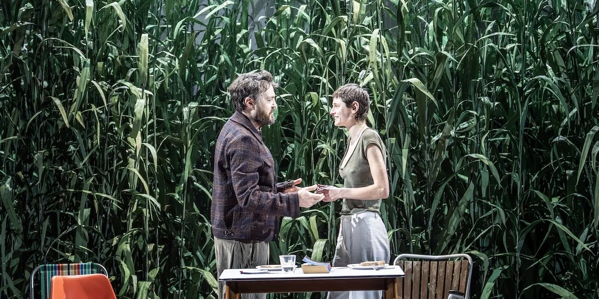 A man and woman exchanging something in a field of tall green plants, with a table and chairs nearby.