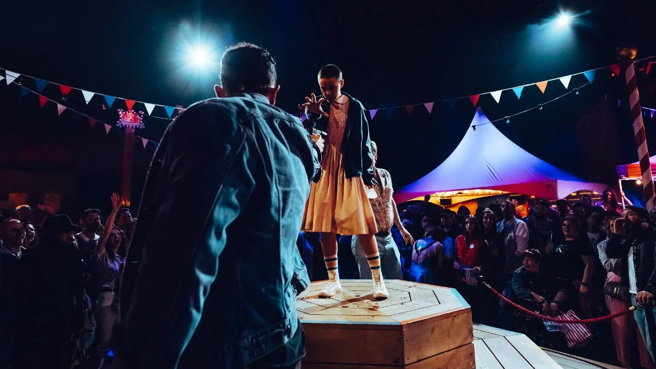 Performers and audience at a circus or carnival event under colorful lights, with a girl standing on a wooden platform in front of a crowd.