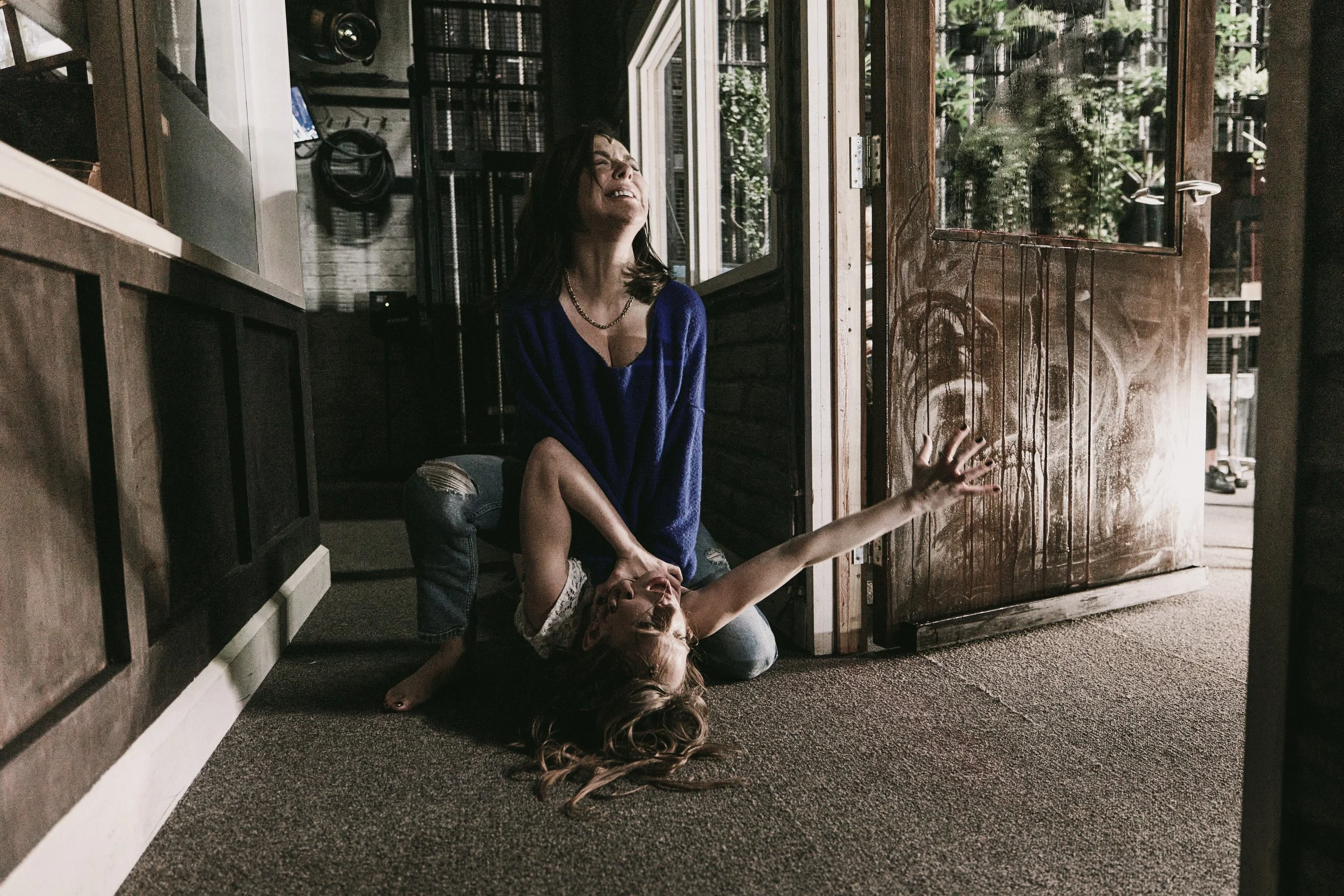 Two women are inside near a glass door and window. One woman is sitting on the floor, with her head tilted back and her eyes closed, appearing distressed. The other woman is kneeling behind her, holding her shoulder and seeming to comfort her. The scene is illuminated by natural light coming through the glass, with greenery visible outside.