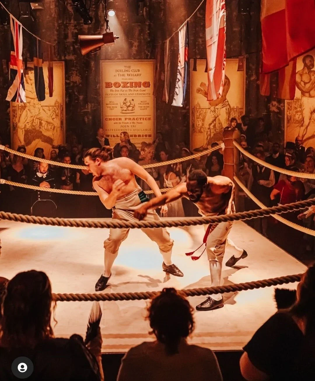 Two shirtless men are fighting inside a boxing ring with ropes, while an audience watches from the background and foreground. The setting has vintage-style boxing posters and flags hanging from the ceiling.