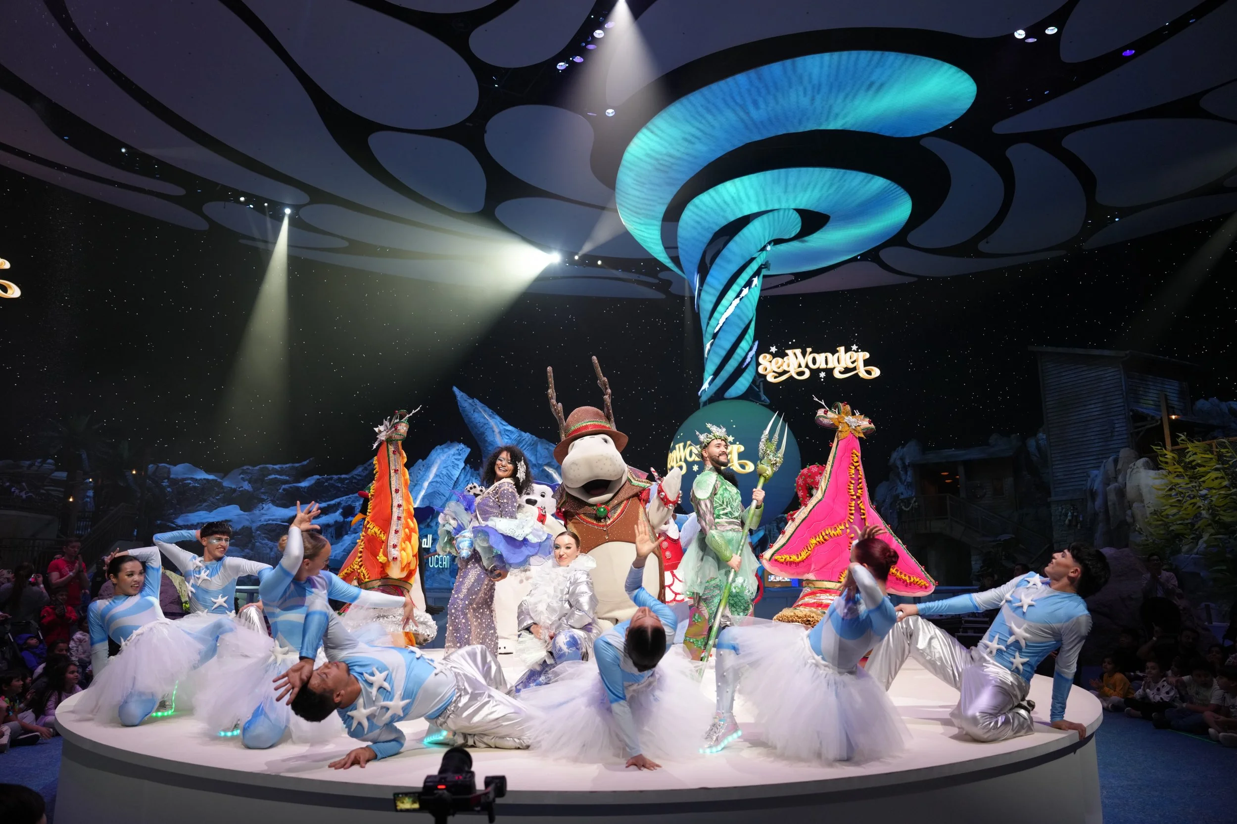 A lively stage performance at SeaWorld with performers dressed as mythical and fantasy characters, including a woman in a purple dress, a man in a reindeer costume, and dancers in blue and white costumes, with a large swirling blue and black ceiling design and the SeaWorld sign in the background.