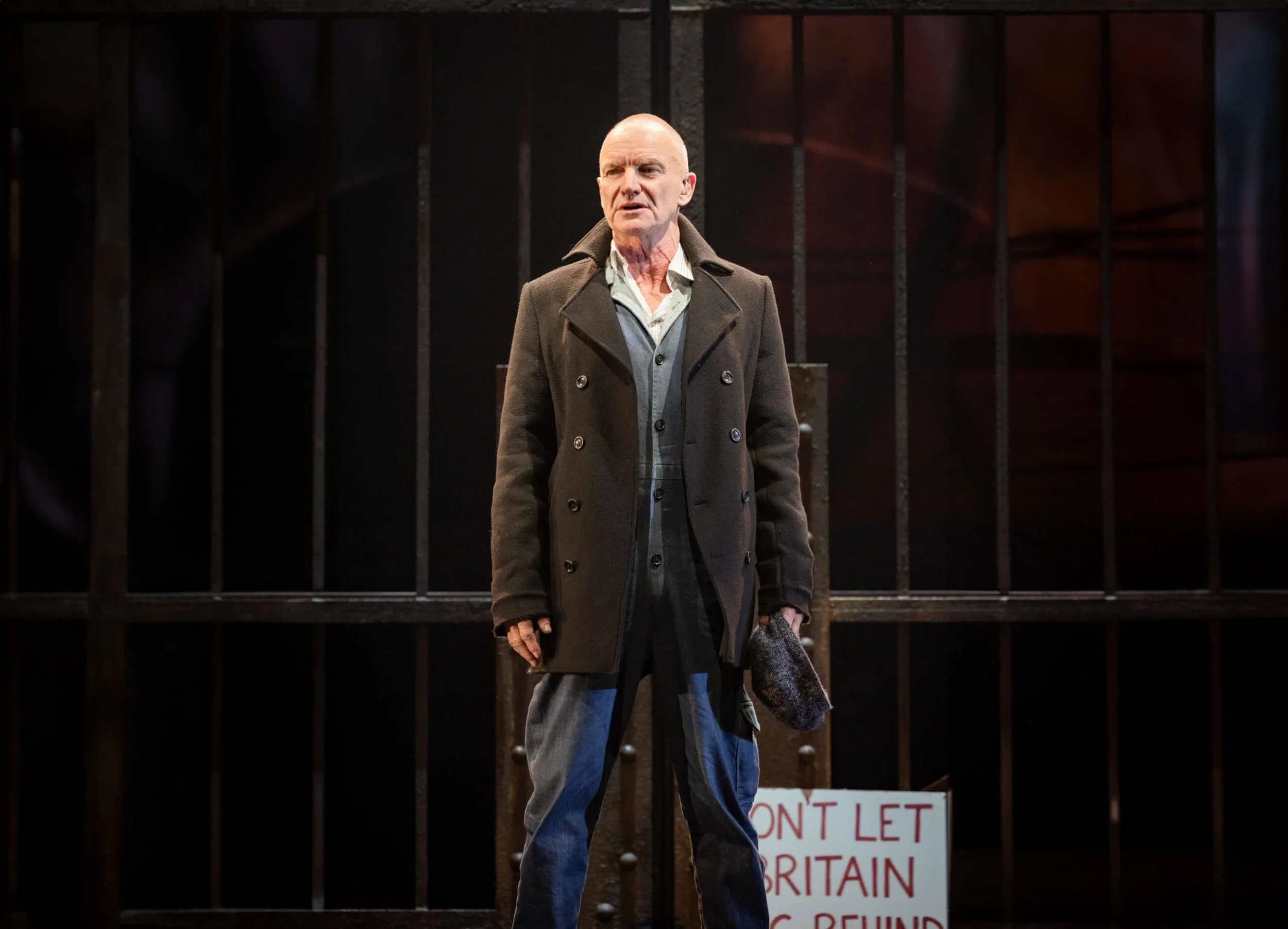 An elderly man with a bald head wearing a dark gray coat over a light-colored shirt and blue jeans, standing in front of a black gate with vertical bars, holding a gray woolen hat in his right hand. There is a partially visible white sign with red text behind him that reads, 'DON'T LET BRITAIN...'