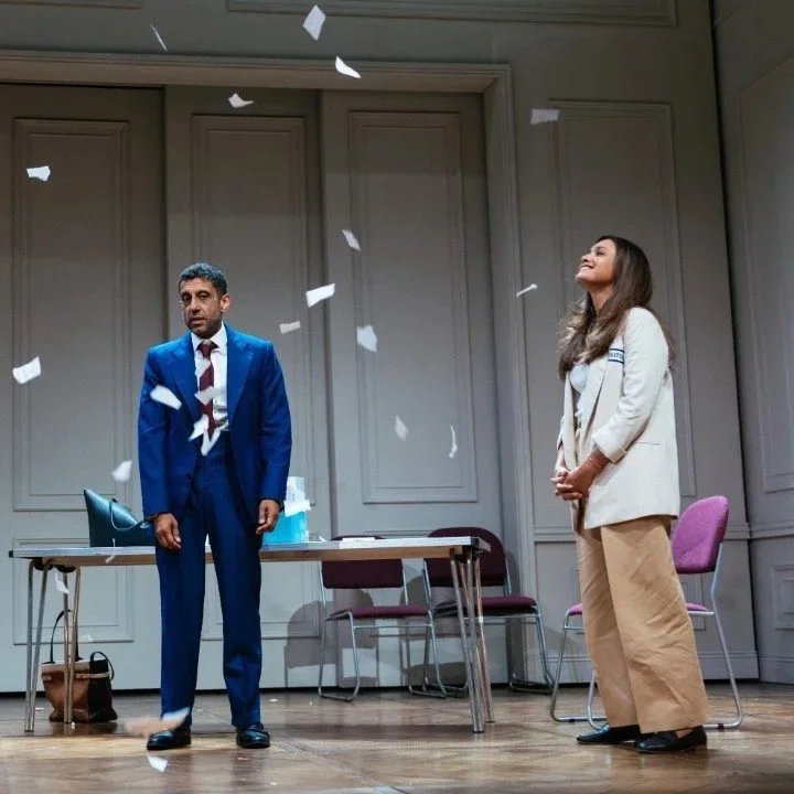 A man in a blue suit and a woman in a beige pantsuit stand on a stage with confetti falling around them, in a formal setting with chairs and a table in the background.