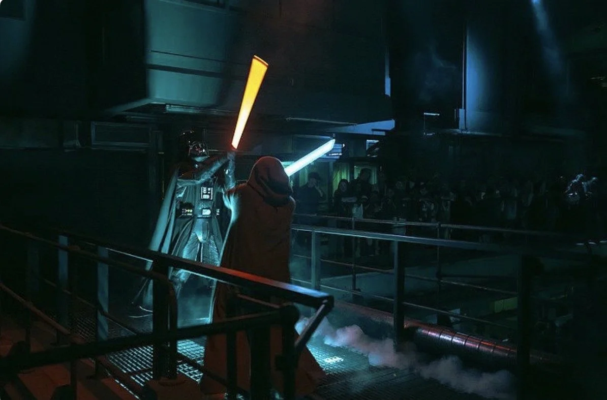 Star Wars cosplay characters with Darth Vader and a Jedi holding lightsabers in a dark, sci-fi themed setting with a crowd in the background.