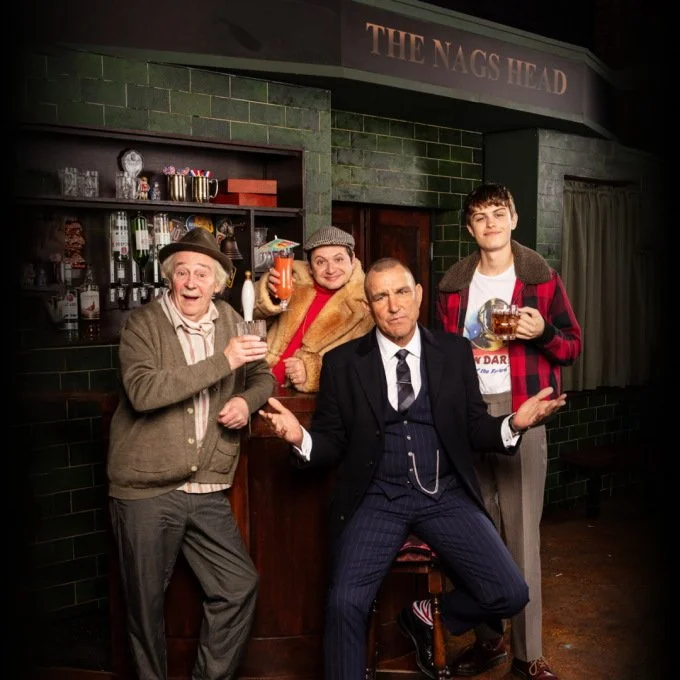 Four men posing inside a pub called 'The Nags Head,' with drinks in hand, smiling and making gestures for the camera.