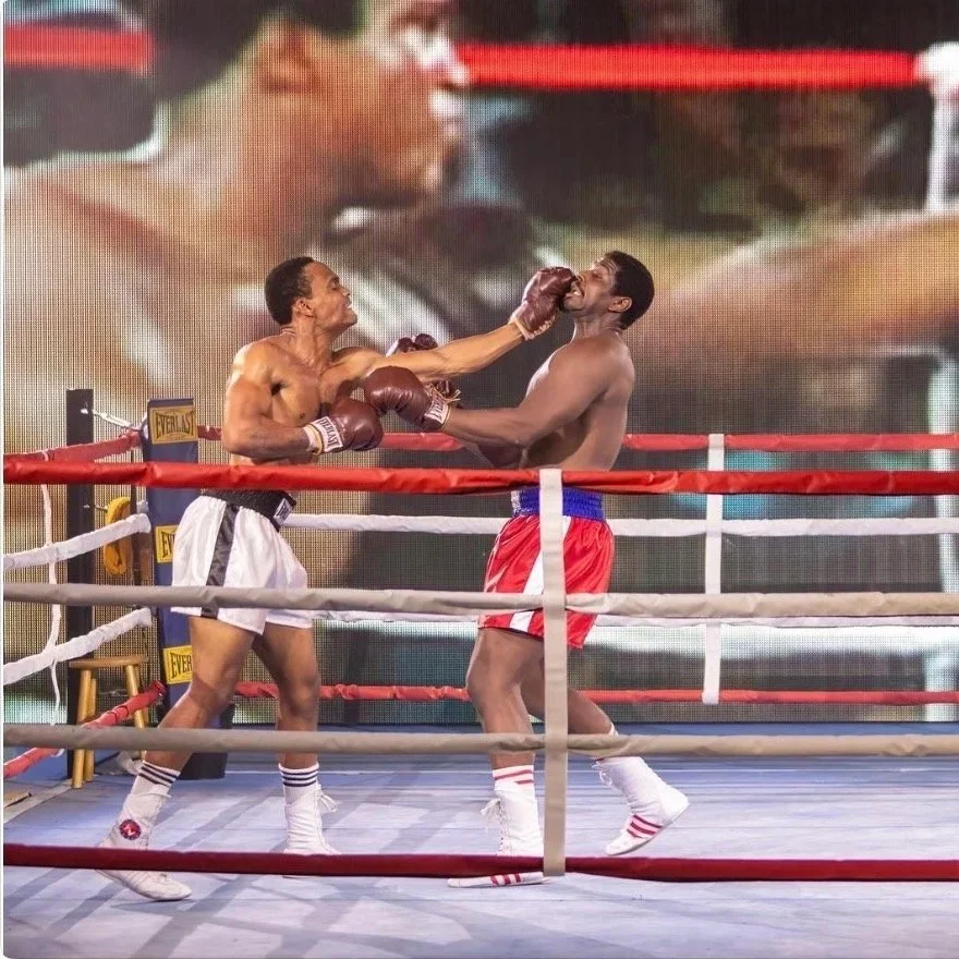 Two shirtless boxers fighting in a boxing ring with red and white ropes, one boxer delivers a punch to the other's face.