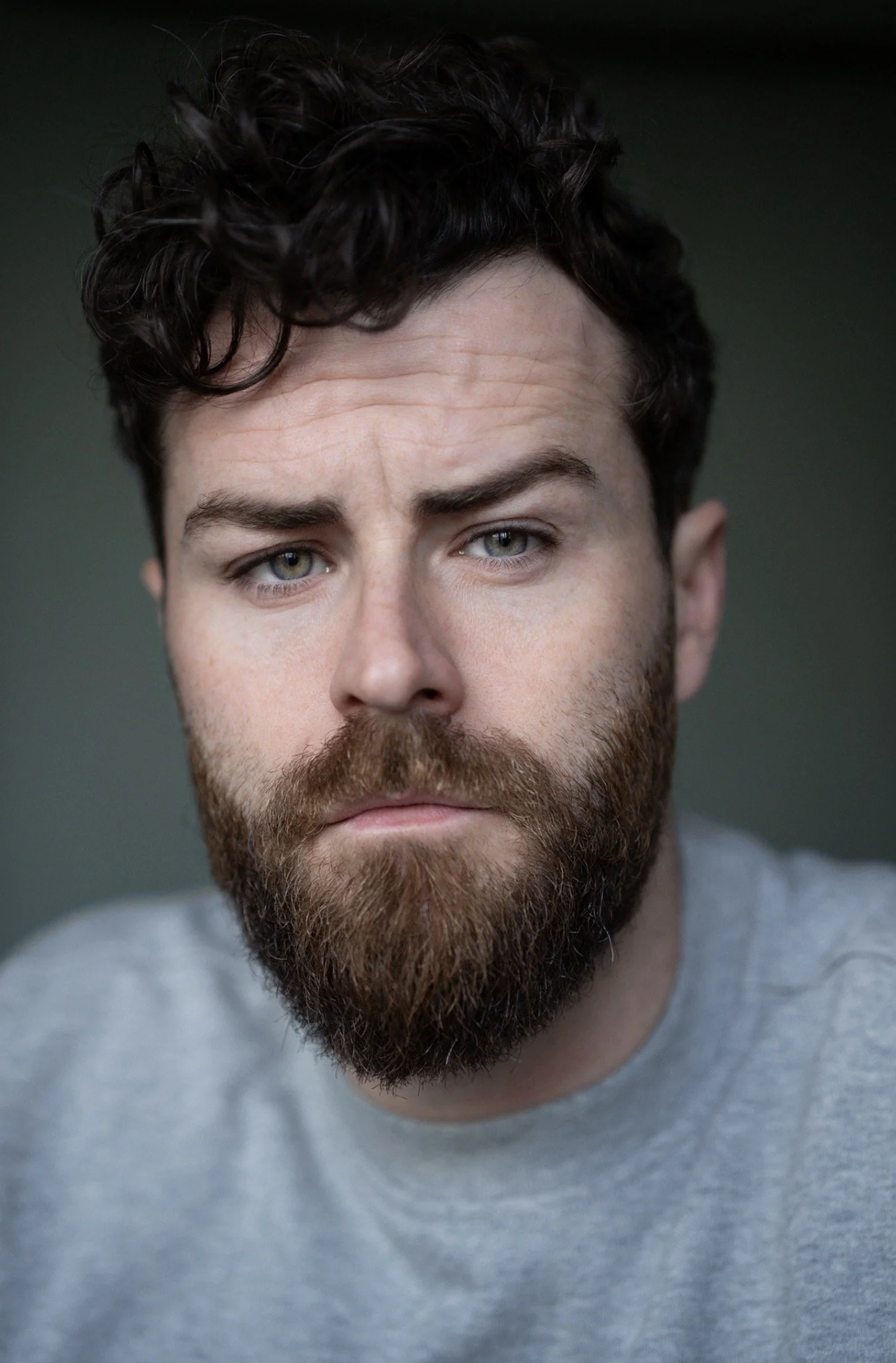 Close-up of a young man with blue eyes, curly dark hair, light skin, and a full beard, wearing a gray T-shirt.