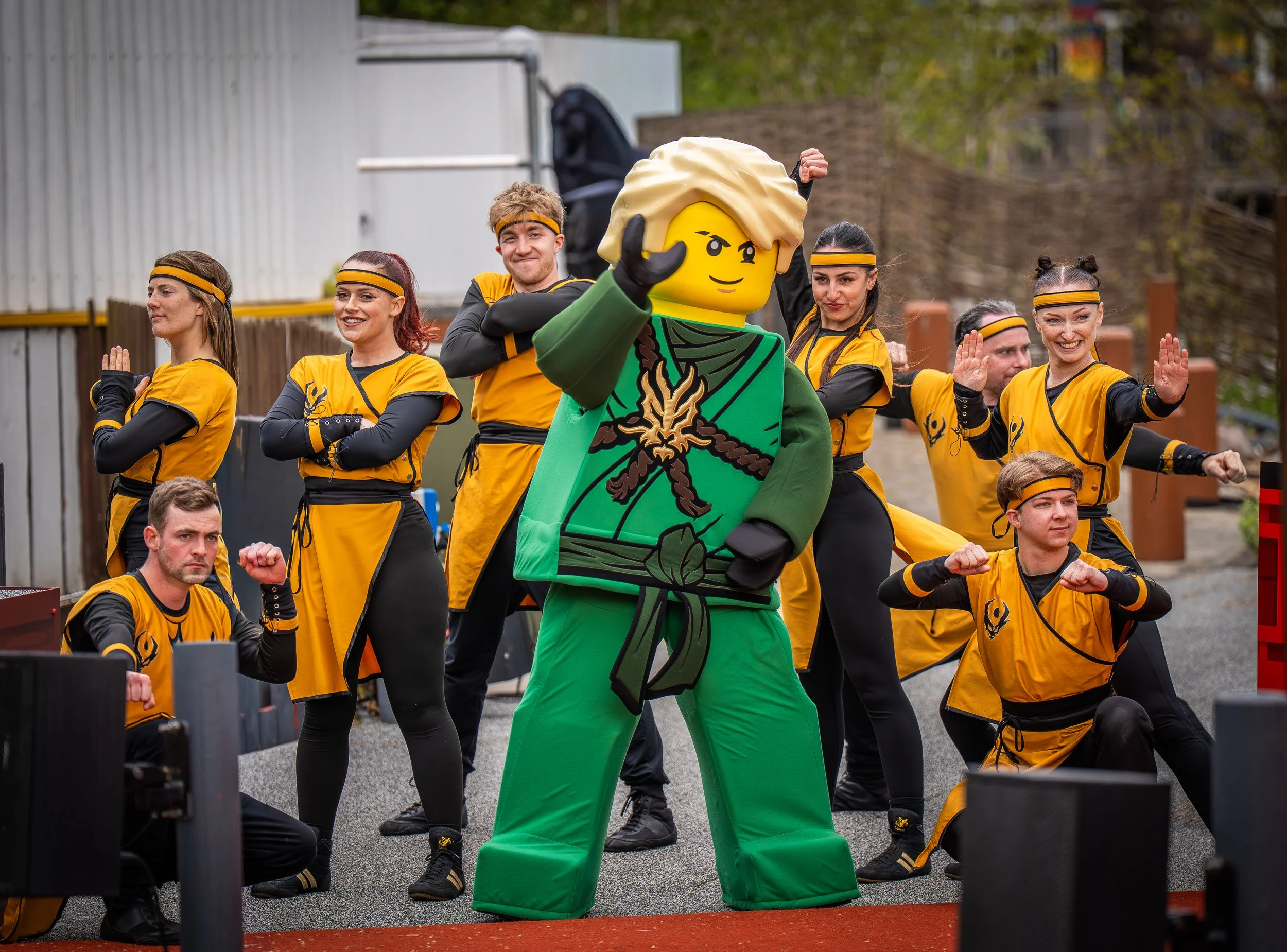 Group of people dressed in yellow and black sports costumes, with some performing martial arts poses, along with a person in a Lego character costume, outdoors in an urban setting.