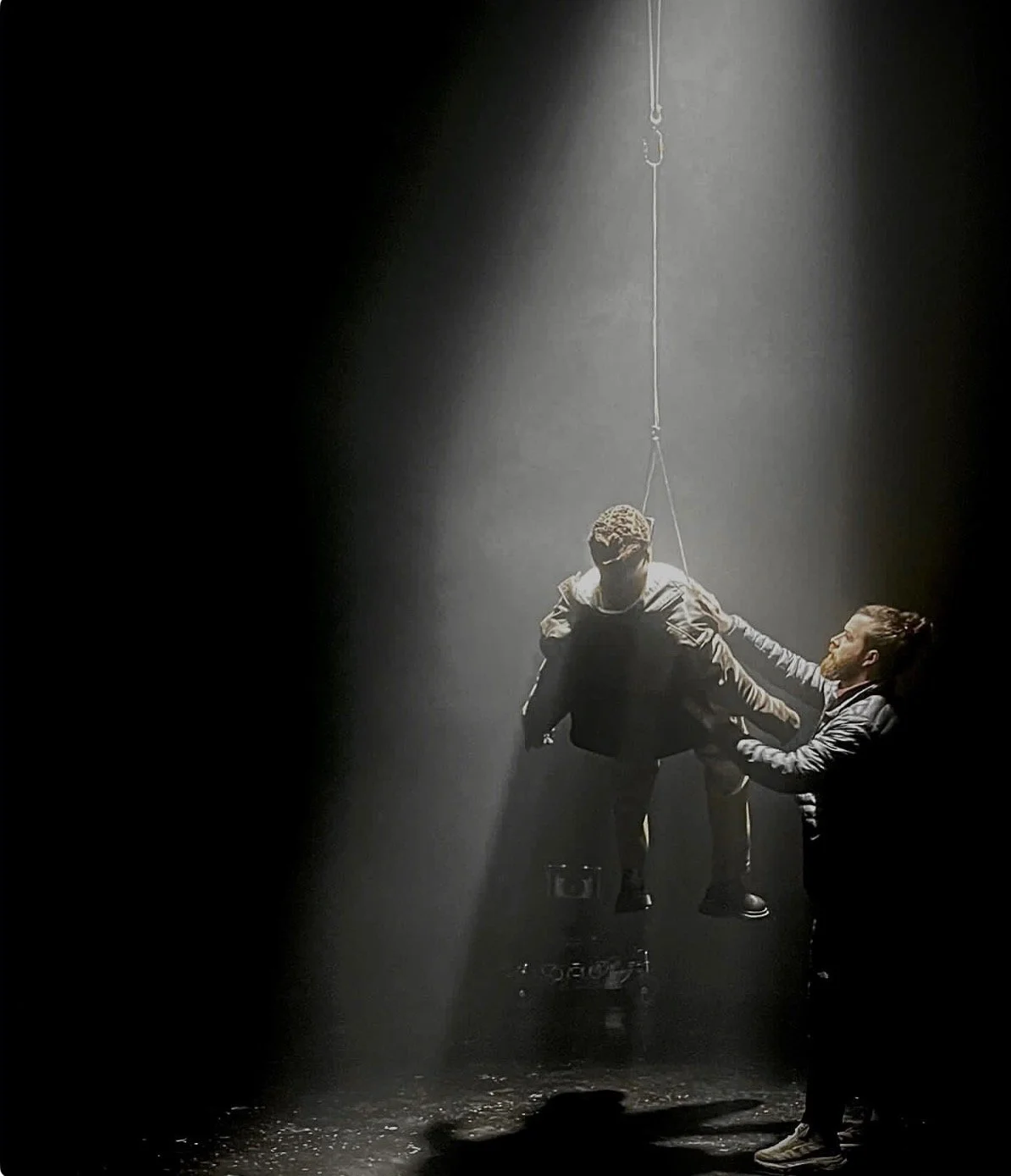 Two men performing an acrobatic stunt on a dark stage with spotlight, one hanging upside down while the other helps.