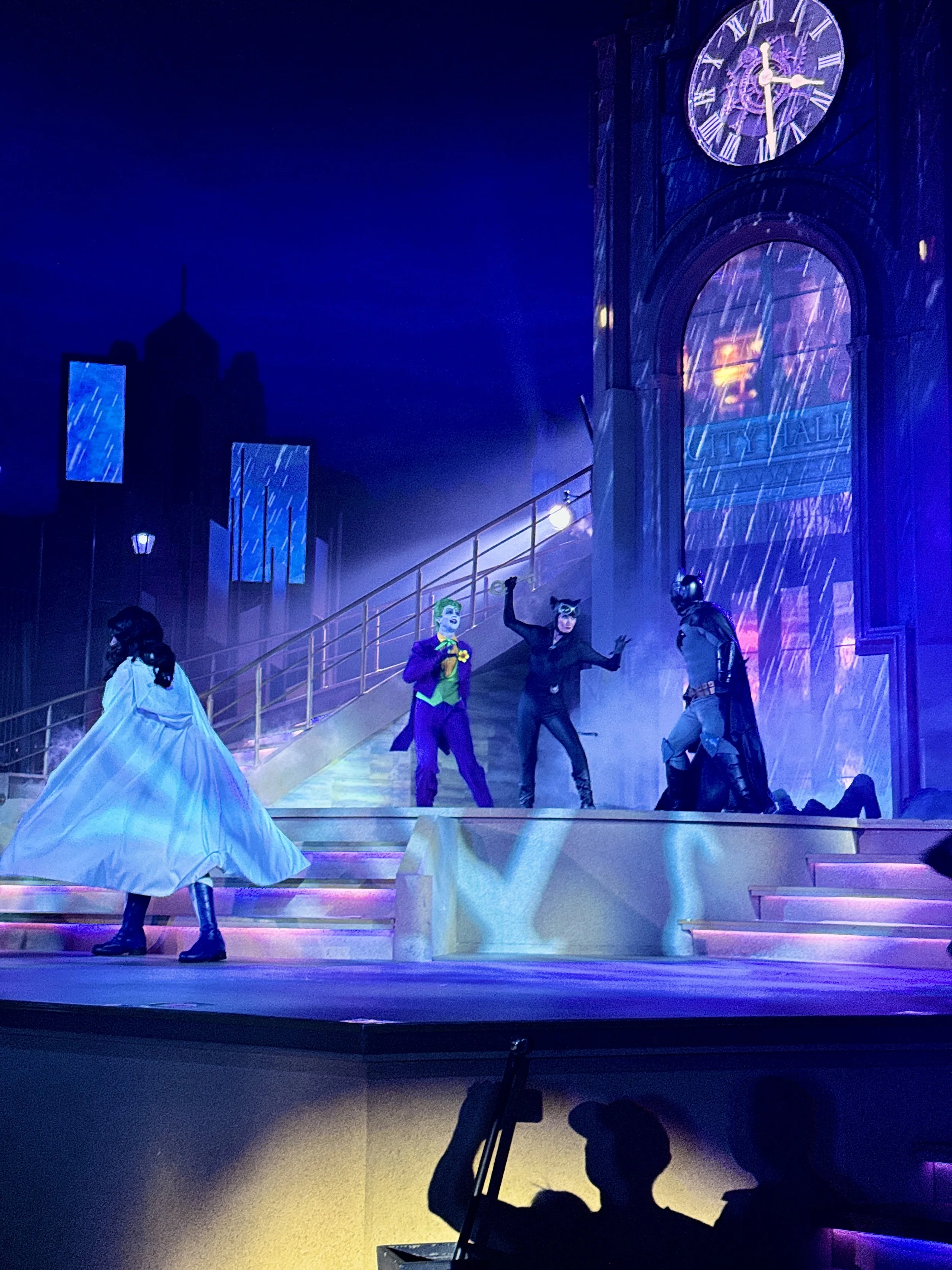 Scene from a theatrical performance featuring characters dressed as the Joker, Catwoman, and Batman, set against a backdrop of a cityscape at night with a large clock and rain effects.